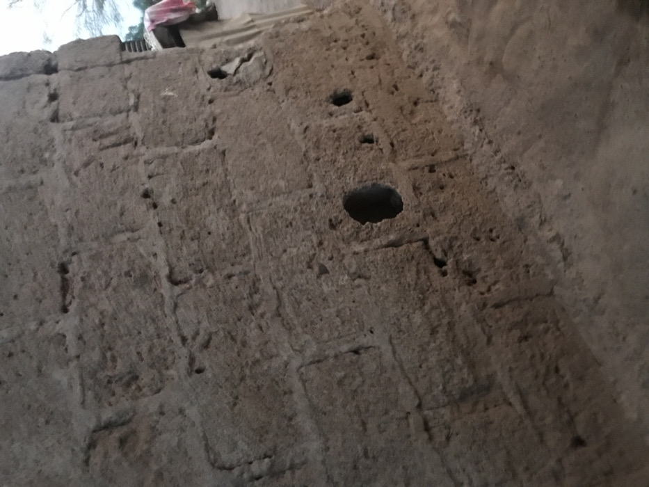 The image presents a close-up, upward-angled view of a weathered wall in Ago-Abo, Nigeria. The wall is constructed from rough, earthy-toned materials, possibly mud bricks or coarse concrete blocks, exhibiting a heavily textured and eroded surface. Visible lines suggest the individual blocks used in its construction.
Several holes of varying sizes are prominent features on the wall's surface. A large, dark, oval-shaped hole is situated in the upper-middle section, with a smaller circular hole above and to its left. Numerous other smaller indentations and imperfections dot the entire wall, suggesting natural wear, degradation, or possibly the remnants of previous fixtures.
At the very top edge of the image, beyond the wall, a bright, diffuse light indicates daytime. A glimpse of distant foliage, likely a tree, is visible against the sky. Additionally, a fragment of what appears to be another structure or a roofline is present, with a piece of reddish-pink fabric draped over it.
The scene is static, devoid of any visible human or animal activity. The rustic construction materials and weathered appearance suggest an older structure within a rural or traditional setting. There is no visible text in the image.
