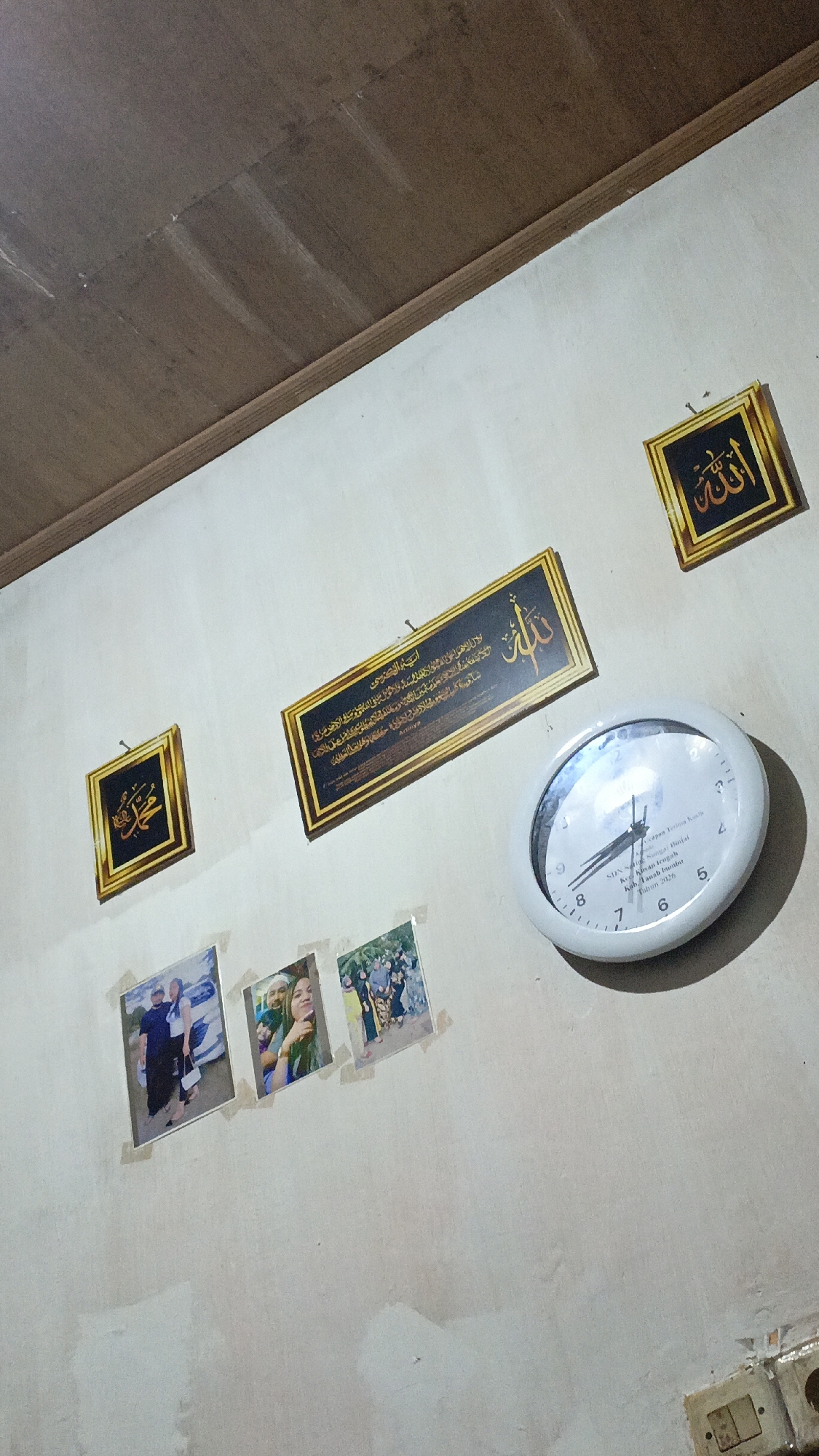 The image depicts a cluttered interior wall in a room located in Pejala, Indonesia. The wall is painted a light, uneven color, with visible water stains and patched areas, suggesting it is in a state of disrepair. A wooden ceiling border runs along the top of the frame.
Several items are affixed to the wall. On the upper right, a framed piece of Islamic calligraphy with a gold border reads "Allah" in Arabic script. To the left of this, a larger, horizontal framed artwork also displays Arabic calligraphy with a gold border. Below these, three framed photographs are taped to the wall. The leftmost photo shows a man and a woman standing near a car. The middle photo is a selfie of two women. The rightmost photo shows a group of people.
A white, circular analog clock is mounted on the wall to the right of the calligraphy pieces. Its face shows Arabic numerals and text that appears to be in Indonesian, including "SDN Sambirejo" and "Tahun 2020." The clock hands indicate approximately 7:45.
The overall impression is of a lived-in space with personal decorations, but also showing signs of wear and tear on the wall.
