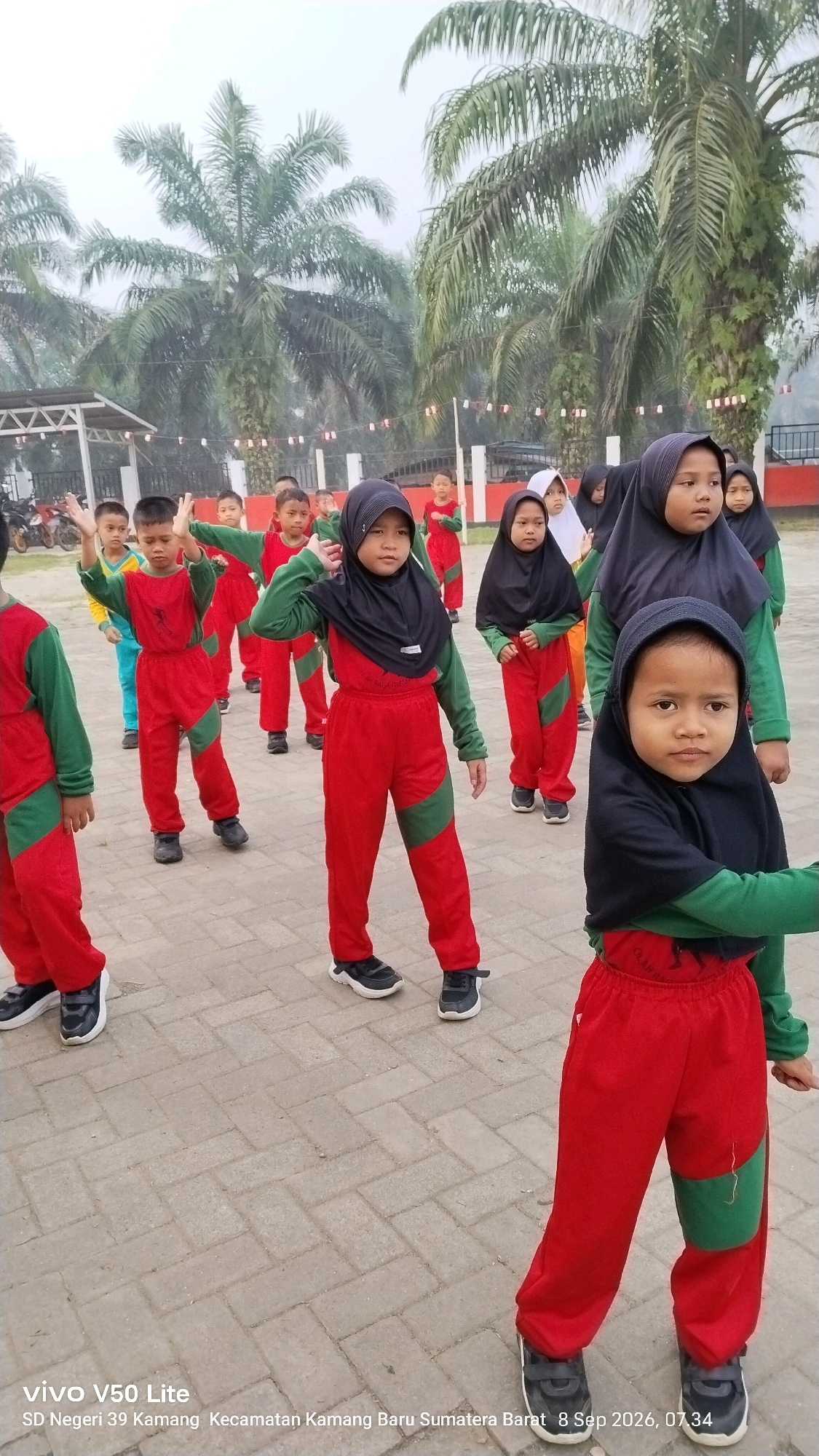 In a schoolyard in Kamang Baru, Sumatra Barat, Indonesia, a group of young students in matching red and green uniforms and black hijabs are engaged in outdoor physical activity. Palm trees loom in the background, suggesting a tropical or subtropical climate. The students, some of whom are facing forward and others in profile, appear to be following instructions, with their arms raised in unison. One student in the foreground, wearing a black hijab and a red and green uniform, is looking directly at the camera. The ground is paved with interlocking bricks. The image also contains a watermark indicating it was taken with a Vivo V50 Lite phone on September 8, 2026, at 07:34.