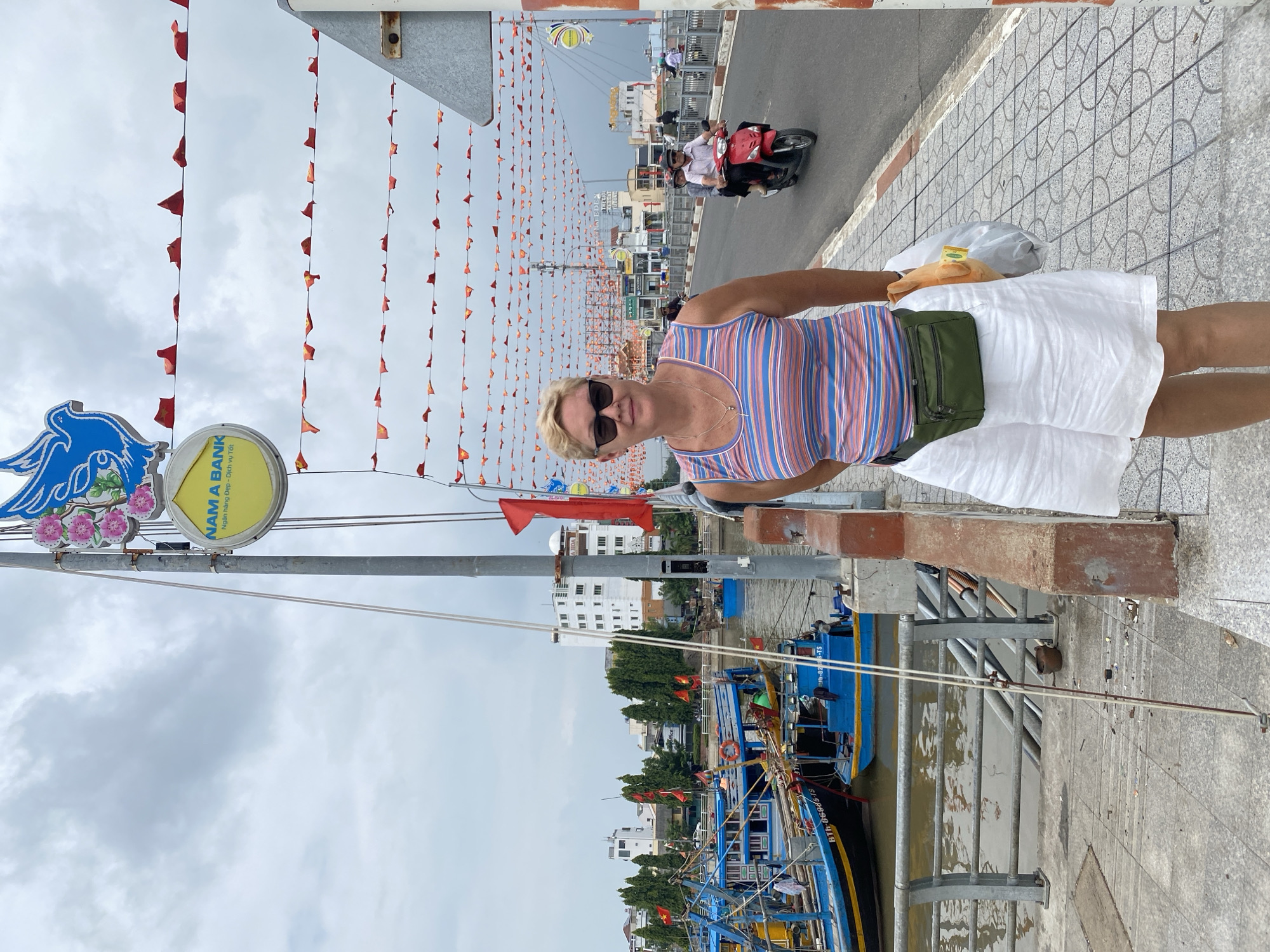 The image shows a woman standing on a sidewalk next to a bridge in Phan Thiết, Vietnam. She is wearing a striped tank top, white shorts, sunglasses, and a green fanny pack. She is holding a plastic bag with a stuffed toy. In the background, there are boats docked in a river, buildings, and people on motorcycles. The sky is overcast, and there are strings of small red flags strung across the bridge. A sign for "NAM A BANK" is visible on a pole. The overall scene depicts a streetscape in a coastal city.