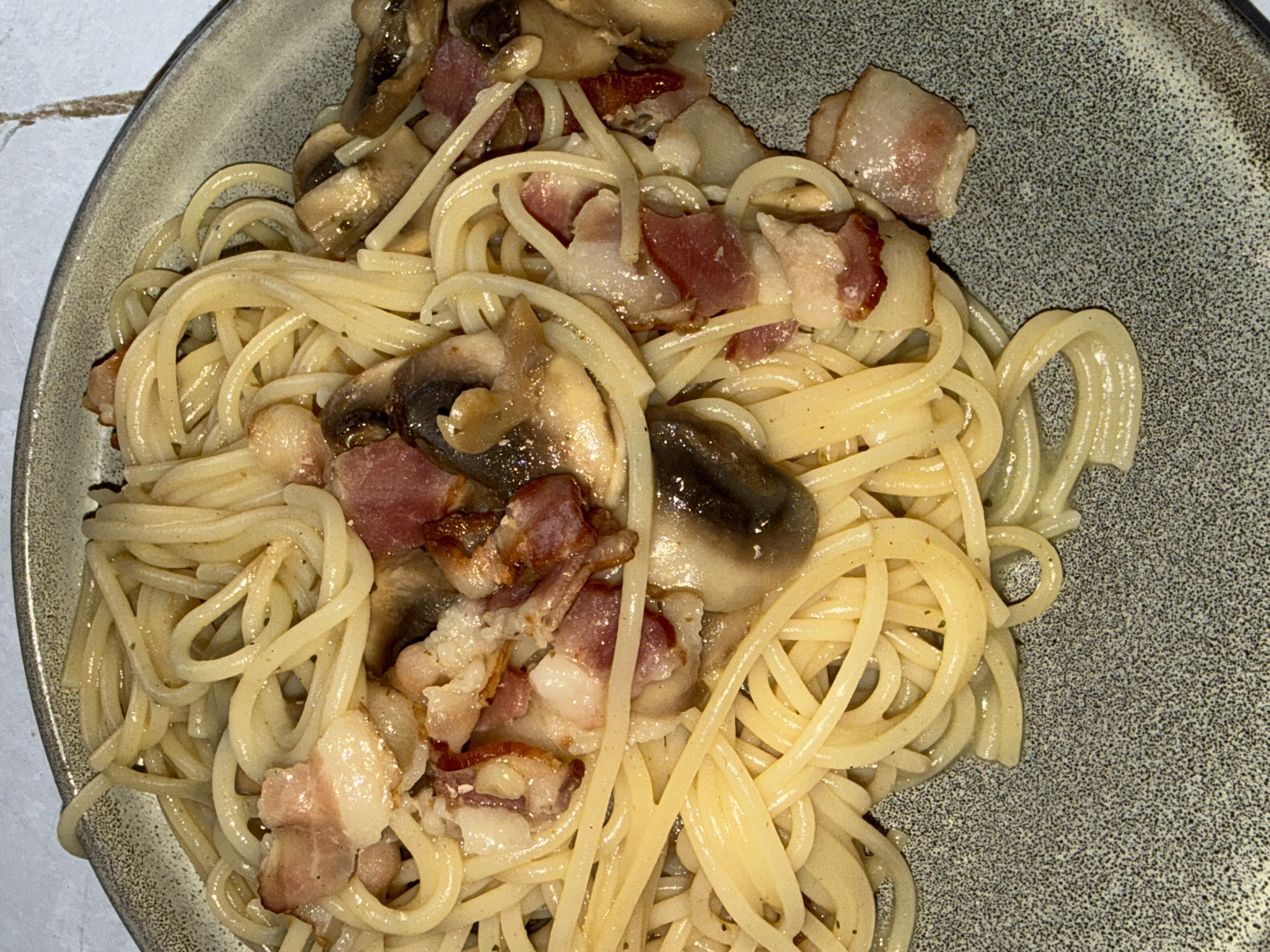 The image shows a close-up of spaghetti with mushrooms and bacon on a grey plate. The spaghetti is light yellow and appears to be coated in a light sauce. Slices of cooked mushrooms and bacon are scattered throughout the dish. The plate is on a white surface. There is no indication of location and there are no people present in the image.