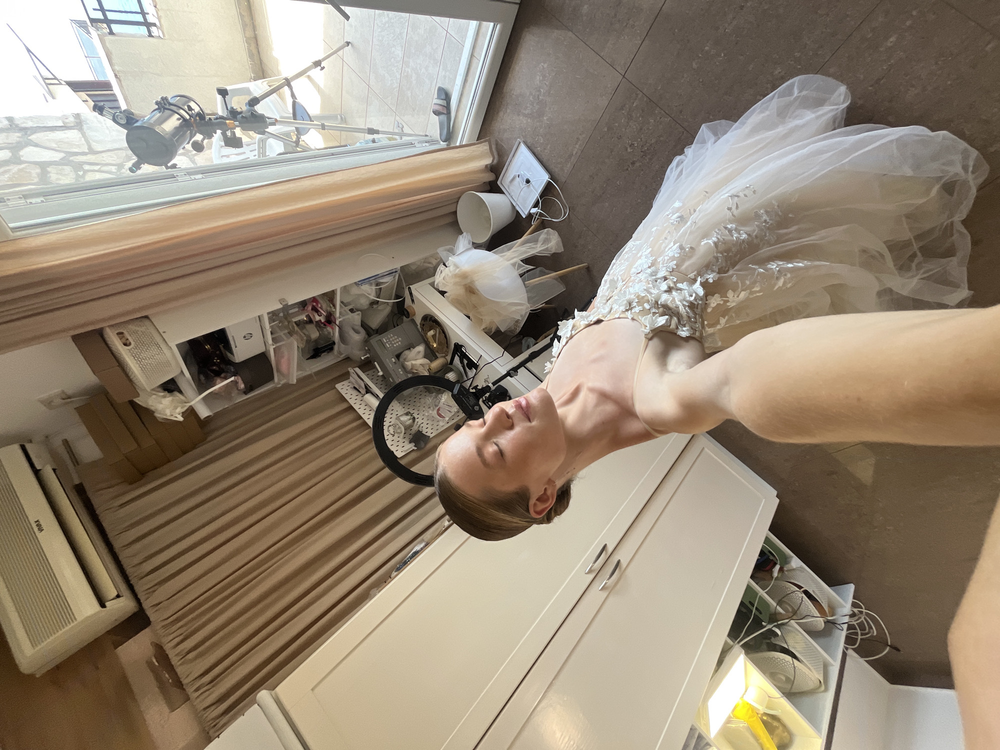 The image depicts a woman wearing a wedding dress, standing in a room that appears to be a dressing room or an area within a home in Budva, Montenegro. The room is furnished with large white cabinets, a beige curtain, and a window leading to an outdoor balcony with a telescope. The woman is positioned in the foreground, with her arm extended as if taking a selfie. She has her eyes closed and her head tilted back, illuminated by sunlight. The wedding dress is detailed with intricate floral embellishments and a flowing tulle skirt. In the background, shelves are visible, containing various items, including a ring light, and what appears to be a sewing machine and fabric scraps, suggesting preparations for a special occasion. The flooring is tiled in a brown hue. The scene conveys a sense of anticipation and beauty.