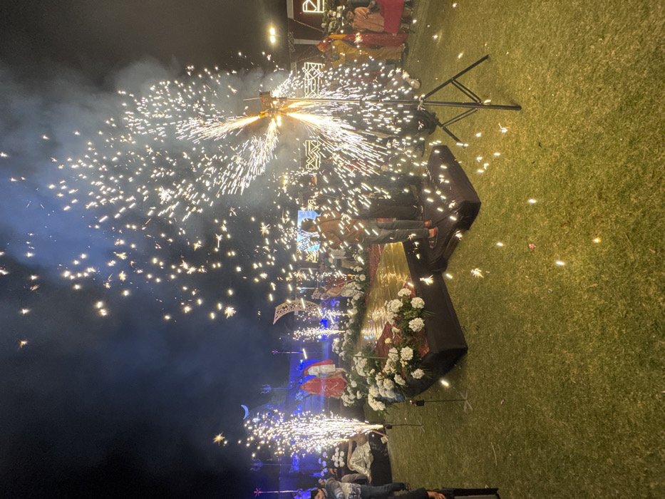 Fireworks explode from a central point, filling the sky with bright sparks. People are gathered near a stage adorned with flowers and a decorative crescent moon. A tripod-like structure holds the fireworks. The scene takes place at night with the foreground of grass. Sparks are visible on the grass and along the edge of the stage. Some individuals appear to be lying down.