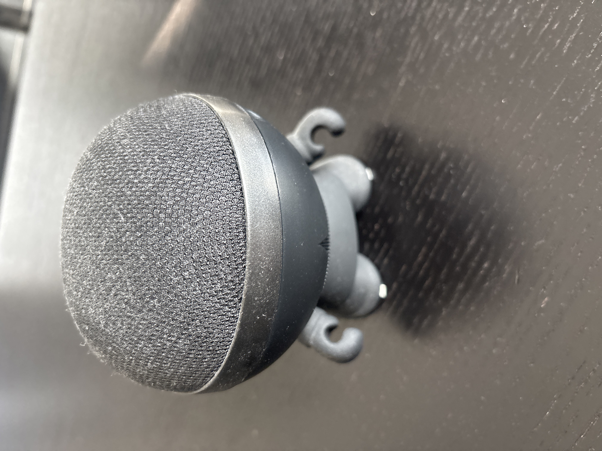 A close-up, eye-level shot shows a small, dark gray, spherical smart speaker with a textured fabric top and a smooth, curved body. It has two small, stubby arms and two short legs with visible white tips, resembling a stylized toy figure. The speaker is positioned on a dark, reflective surface that appears to be a wooden table with visible grain and a subtle sheen. The lighting creates a soft shadow of the speaker on the surface. The background is out of focus, showing a lighter, indistinct area. There are no people or animals present, and the scene is static. This image was captured in Dubai, United Arab Emirates.