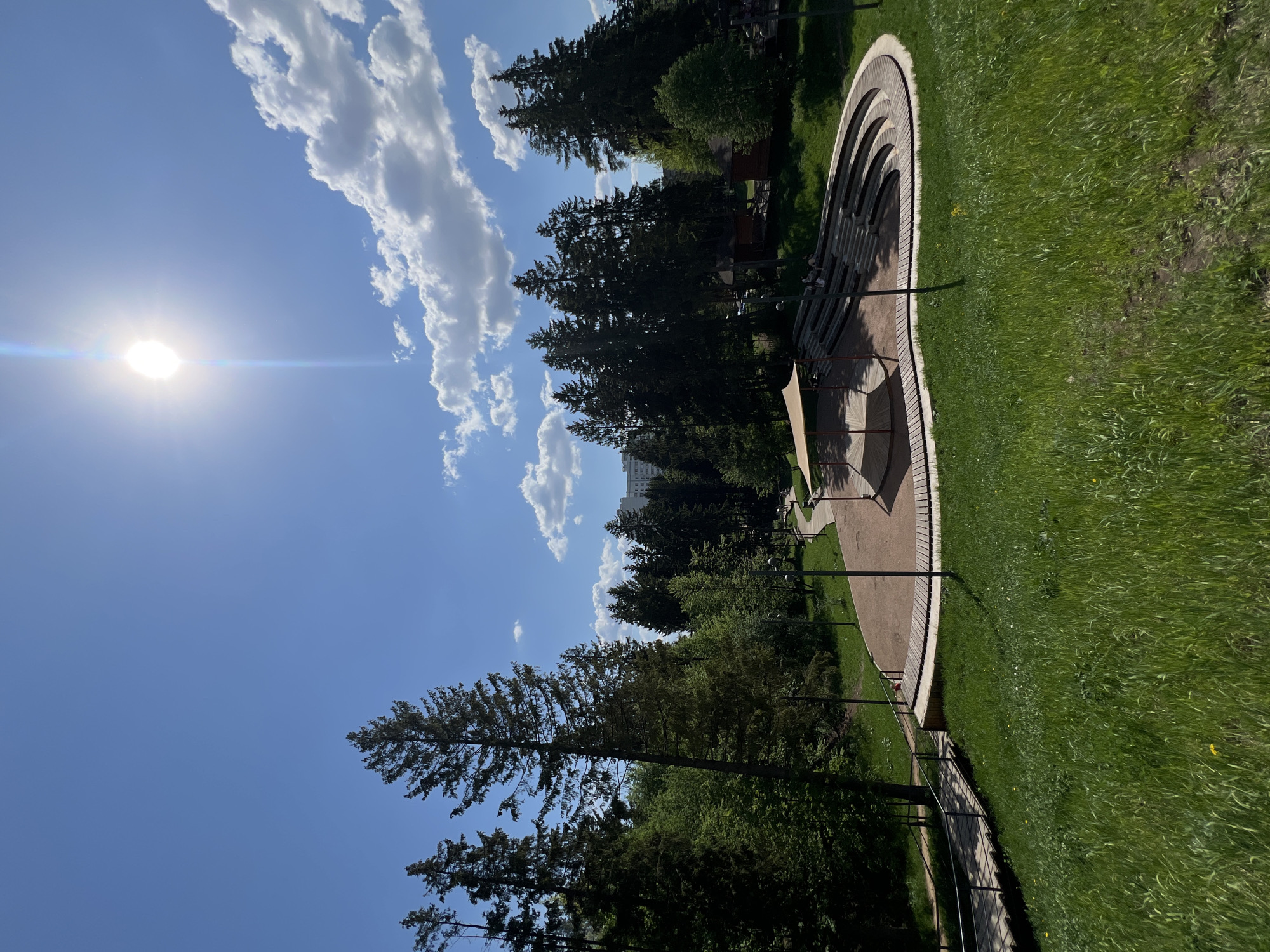 The image depicts a sunny day in a park setting. The sky is blue with scattered white clouds and the sun. A group of trees stands in the middle ground, surrounding an elevated amphitheater-style structure made of light brown material, with multiple rows of seating. A paved pathway with metal railings runs parallel to the amphitheater. The ground is covered in green grass. The photograph was taken from a high vantage point and rotated ninety degrees. It is located in Izhevsk, Russia.