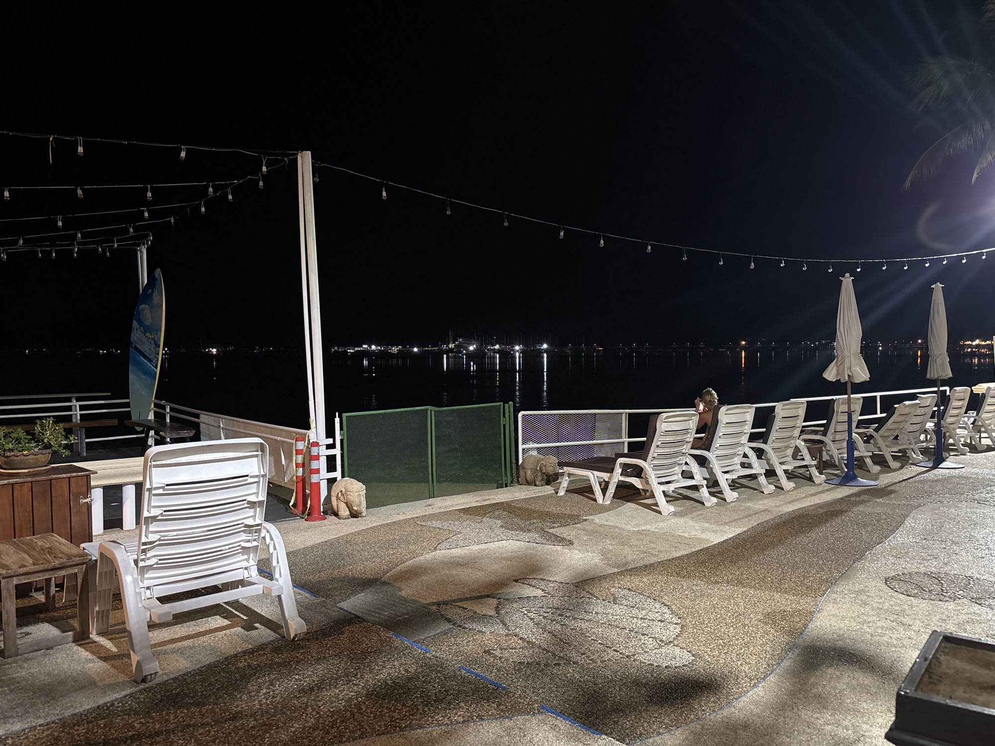 A nighttime outdoor scene in Chalong, Thailand, shows a waterfront promenade with several empty lounge chairs and umbrellas. String lights hang overhead, casting a dim glow. A person is seated in one of the lounge chairs, facing away from the camera. In the background, the calm water reflects the lights of distant boats and buildings. The promenade is paved with a textured surface, and decorative patterns are visible on the ground.