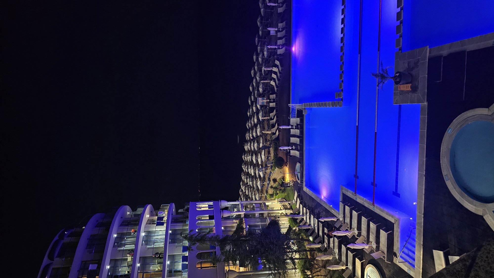 The image depicts a night scene at a resort or hotel in Varna, Bulgaria, likely situated on the coast. A large, modern building with rounded balconies and illuminated windows dominates the left side of the frame. The building appears to be several stories high. In front of the building, a vast expanse of beach chairs and umbrellas is visible, neatly arranged. Adjacent to the beach is a brightly lit, multi-level swimming pool, glowing with a vibrant blue light. The pool features lane markers and illuminated edges. A smaller, circular jacuzzi-like pool is located in the foreground to the left. Beyond the beach and pool area, the dark expanse of the sea is visible under the night sky, with distant lights suggesting a coastline or other structures. There are no visible people in the immediate foreground or performing any specific actions, though some figures can be seen on the balconies of the building. The overall impression is one of a luxurious, well-maintained resort at nighttime.