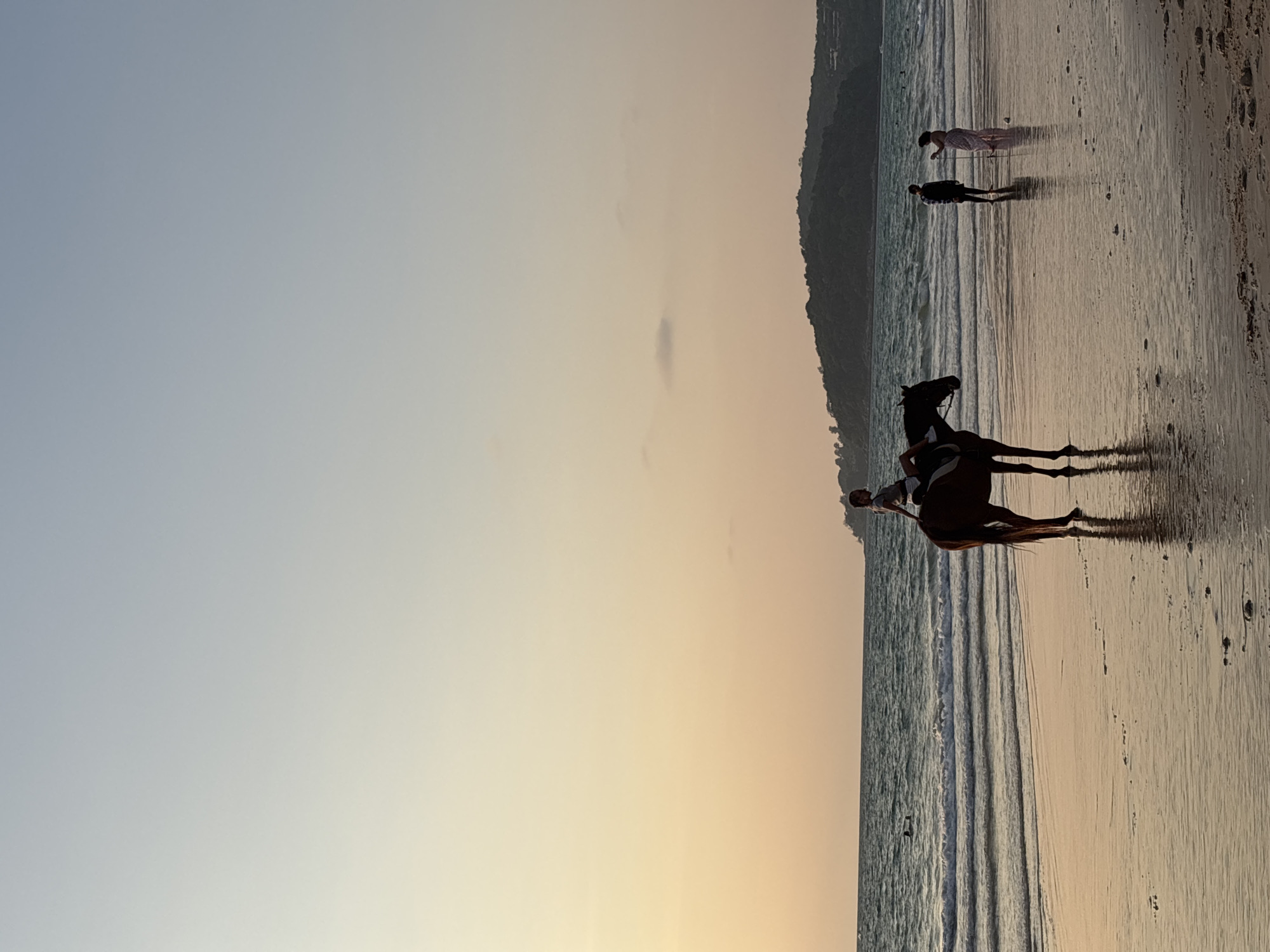 A horse and rider gallop on Bang Tao Beach in Choeng Thale, Thailand, at sunset. The scene includes two other individuals wading in the water behind them. A hillside with foliage is visible in the background, and the sky is a pale gradient of blue and orange.