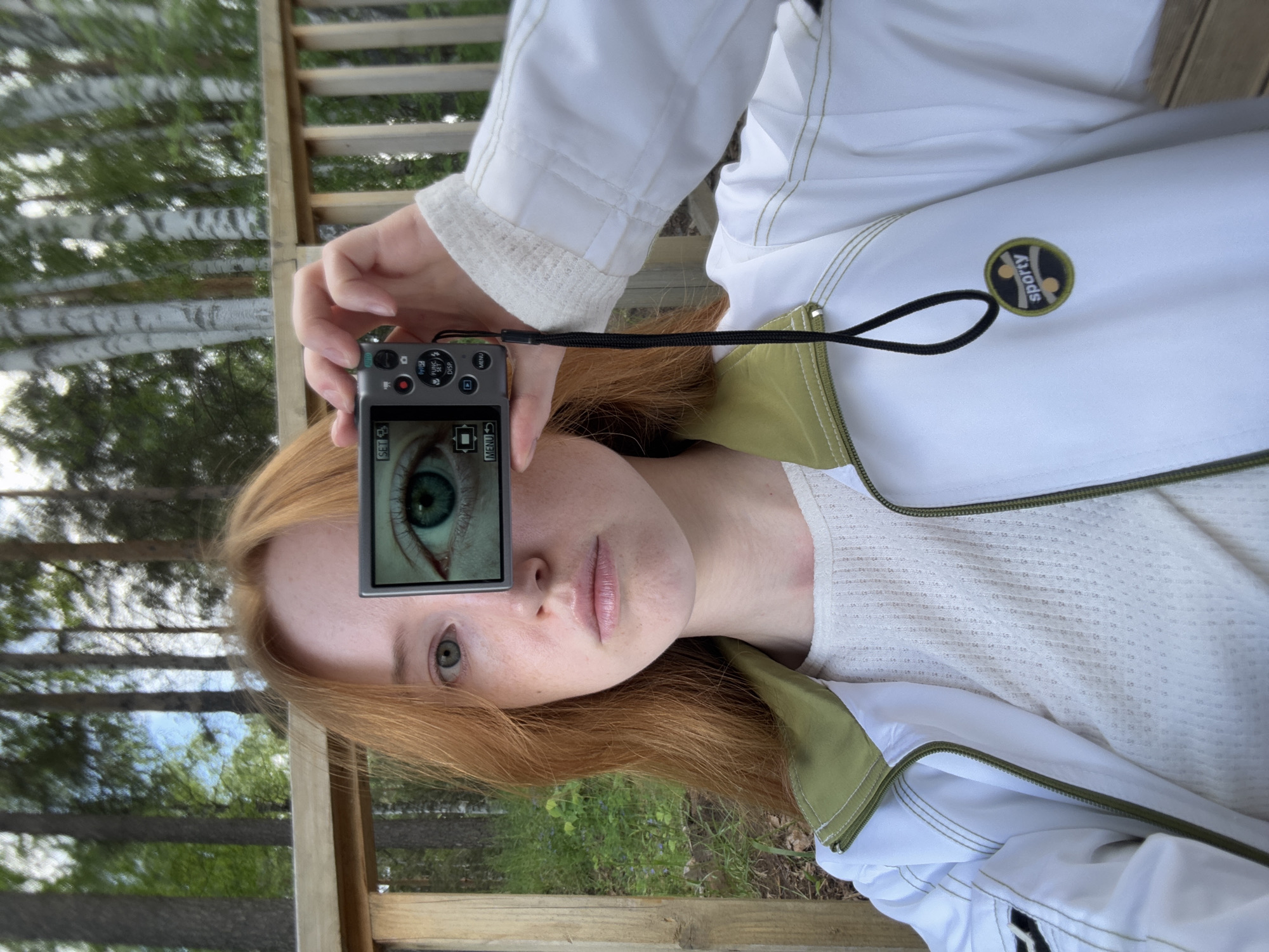 A young, light-skinned person with red hair is holding a gray digital camera up to their face. The camera screen shows a magnified view of one green eye. The person is wearing a white jacket with green accents and a white textured shirt. They appear to be outdoors on a wooden deck or porch, as a wooden railing is visible. Through the railing, trees are visible in the background. The location in Chaikovsky, Russia cannot be verified from the image.