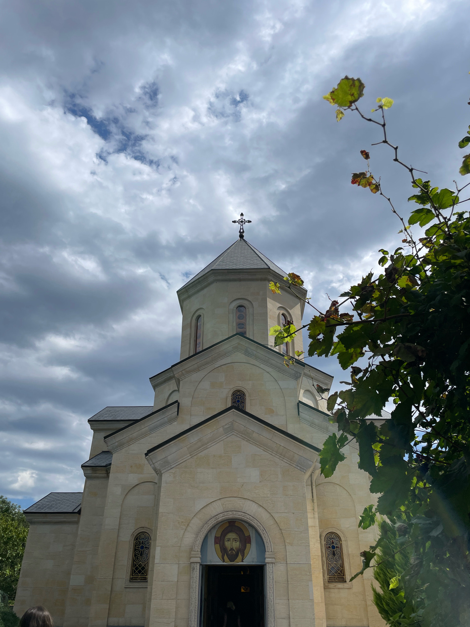 The image shows a light-colored stone church with a cross atop its dome. The church has arched windows with decorative grilles and an icon of Jesus above the entrance. In the foreground, a tree with green and reddish leaves partially obscures the view of the church. The sky is partly cloudy with dark gray clouds. This church is located in Saguramo, Georgia.