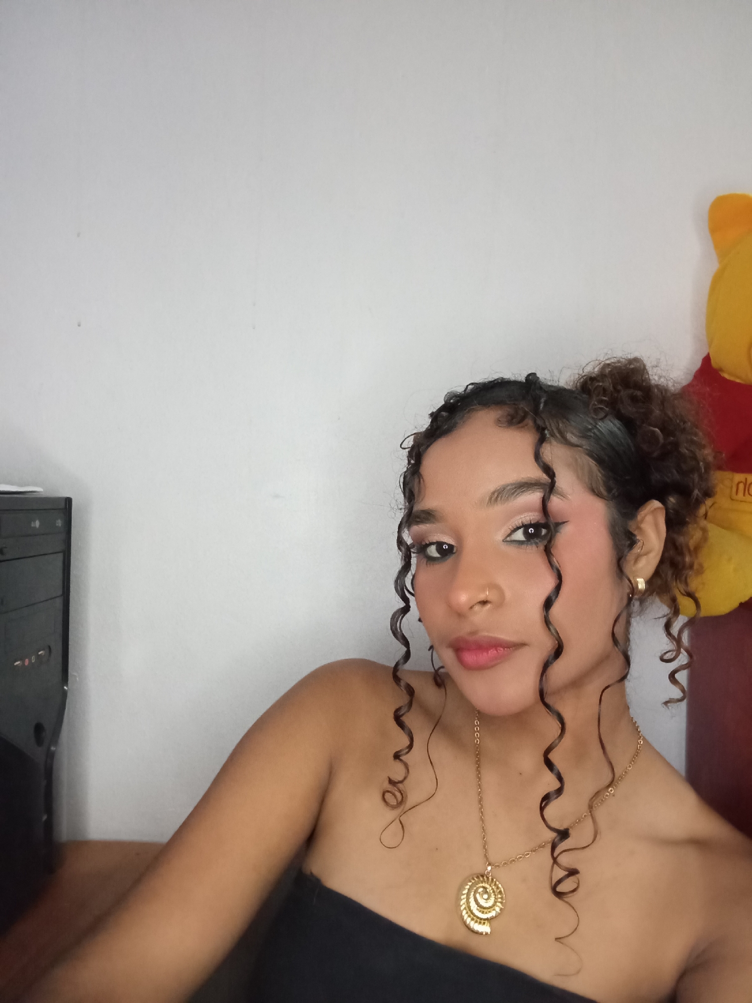The image presents a close-up indoor portrait of a woman in Juan Griego, Venezuela. The subject, a woman with medium skin tone and dark, curly hair, looks directly at the camera with a neutral expression. Her hair is styled with individual curls framing her face, while the remainder is pulled back. She wears a strapless black garment. Visible accessories include a gold hoop nose ring in her right nostril, small gold hoop earrings, and a gold chain necklace featuring a spiral shell-shaped pendant. Her face displays applied makeup, including winged eyeliner, eyeshadow, blush, and pink lipstick. The background consists of a plain, light-colored wall. To the left, the top portion of a black computer tower is visible on a wooden surface. To the right, a segment of a yellow plush toy, featuring a red garment element, is partially visible.