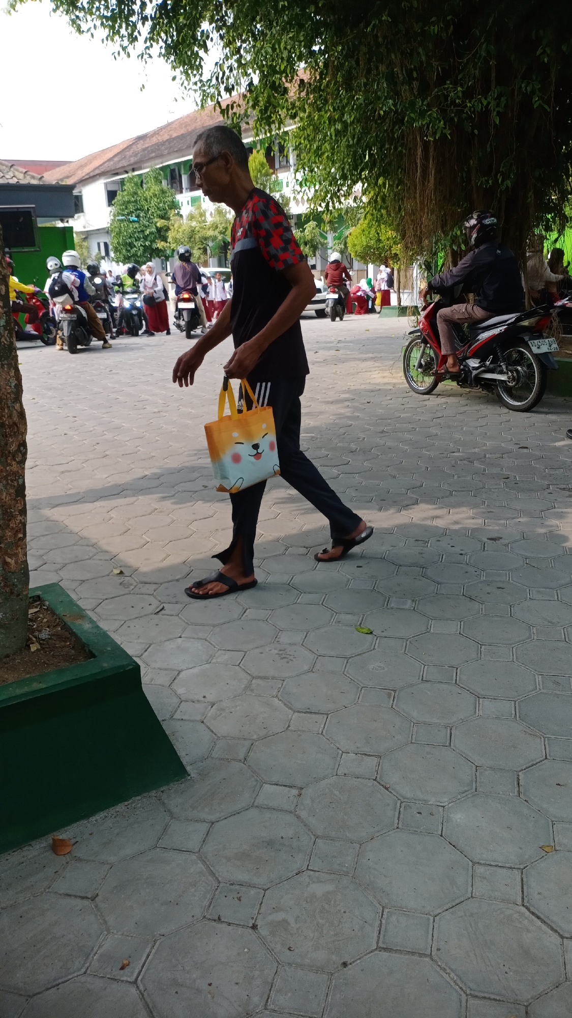 An elderly man with glasses is walking to the right, carrying a yellow tote bag with a Shiba Inu dog graphic. He is wearing a black t-shirt with a red and black geometric pattern on the sleeves and dark pants. He is wearing black sandals. The man is walking on a paved walkway made of hexagonal and square concrete tiles. Behind him, in the background, is a school building. Several people on motorcycles are visible in front of the school building. The area is outdoors, with trees and foliage visible, suggesting a sunny day.
