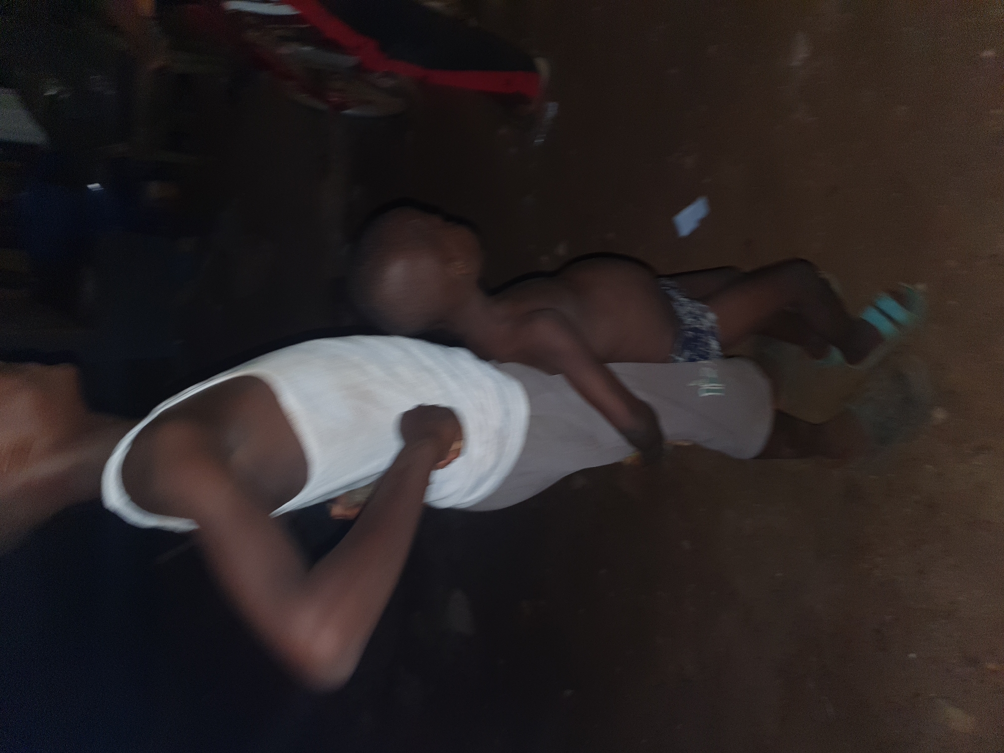 The image captures an intimate moment between an adult and a child in a dimly lit, possibly nighttime or indoor setting. The photograph is taken from a high angle, looking down.
The primary subjects are two individuals, both appearing to have dark skin tones. On the left, an adult is partially visible, wearing a white tank top. A young child, clad in patterned shorts and light blue or green sandals, is lying horizontally across the adult's stomach or lap. The child's head is turned away from the camera, and their body appears relaxed, suggesting comfort or rest.
The background is predominantly dark, indicating poor lighting. The floor appears to be made of dirt or dark earth, suggesting a rustic or informal environment. Several small, light-colored objects, possibly debris or paper, are scattered on the ground near the child's feet. In the upper part of the image, a blurry red and black object is faintly visible.
The overall scene conveys a quiet and possibly tender interaction between the two individuals. The low light conditions contribute to a sense of privacy or late hours. No discernible text is visible in the image. Given the context of Idah, Nigeria, the setting's simple nature and dim lighting could be characteristic of some local environments.