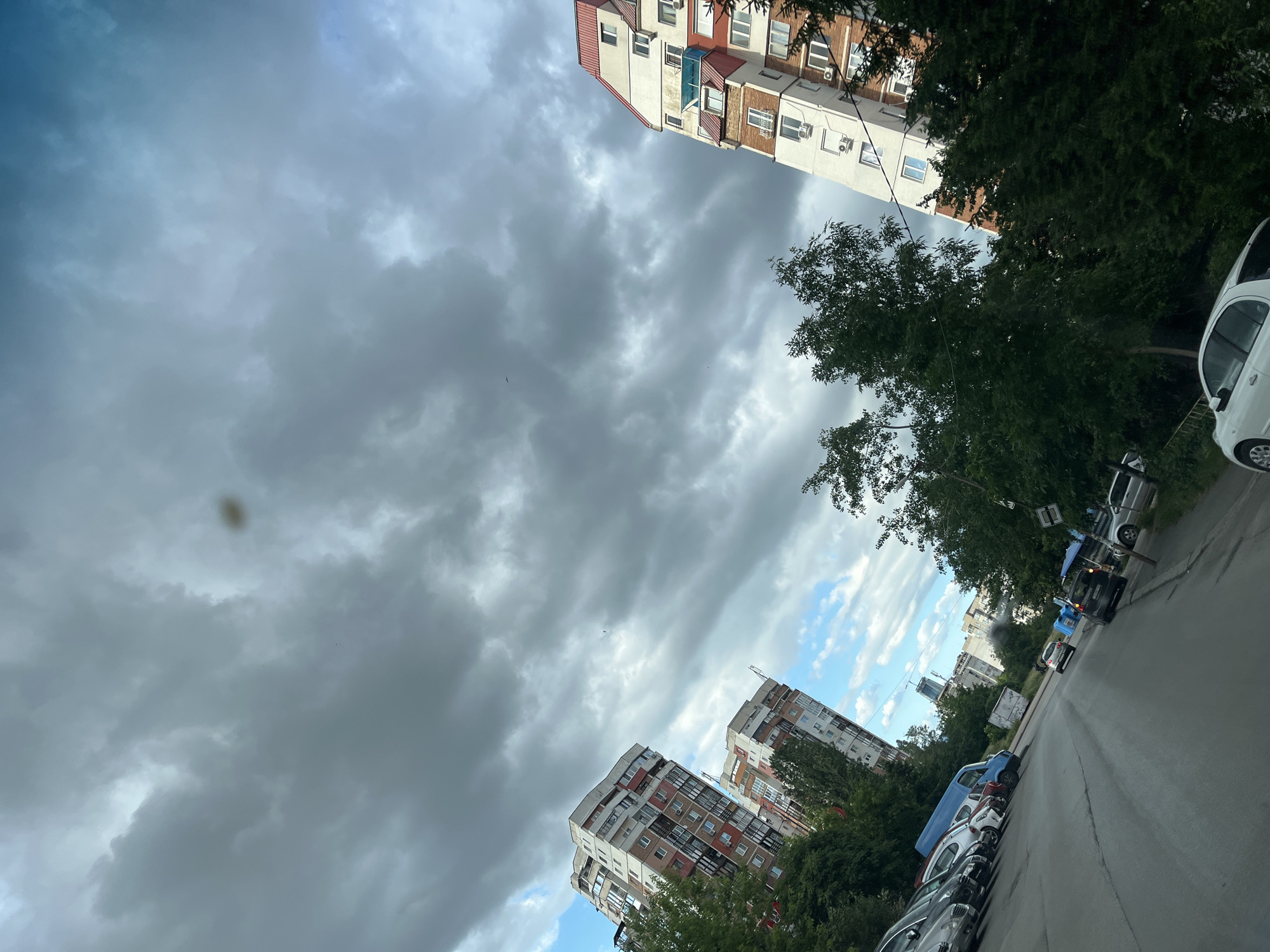 An eye-level shot, looking up from a street, captures a cloudy sky over an urban environment. Tall residential buildings with white and reddish-brown facades dominate the skyline, angled due to the camera's perspective. Lush green trees flank the buildings and line the street, obscuring some of the lower portions of the structures. Parked cars are visible along the side of the asphalt road, and a few vehicles are in motion. The sky is a mix of dark, heavy clouds and patches of lighter blue, suggesting an overcast or potentially stormy day. A small, blurry brown spot is visible in the upper portion of the sky.