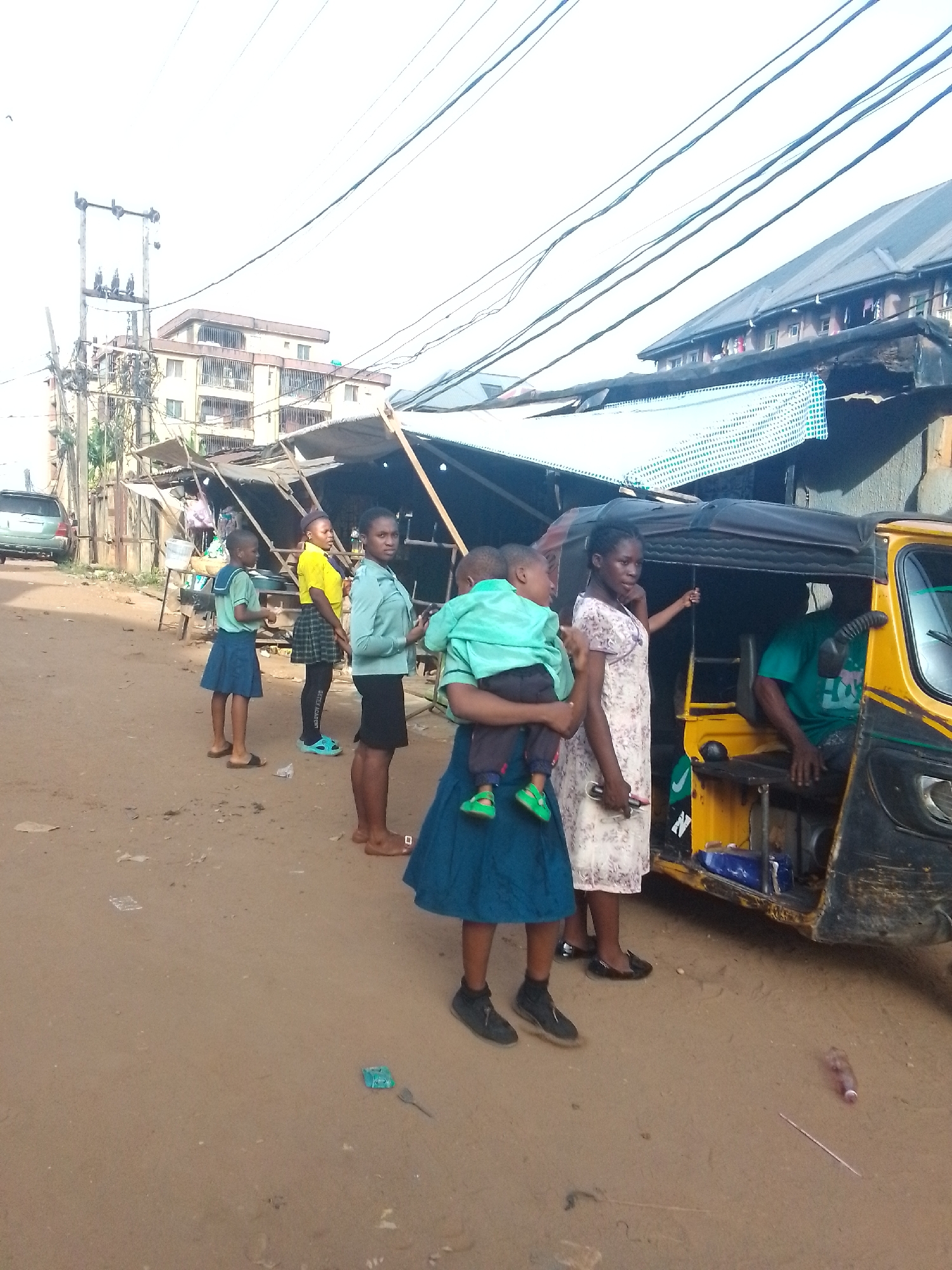 The image shows a street scene in Nkpor, Nigeria. A yellow and black auto-rickshaw (tricycle) is parked on a dirt road. Several people are standing near the vehicle. A woman in a blue dress is holding a small child dressed in green. Another woman in a floral dress is standing beside them, seemingly interacting with the auto-rickshaw driver. Other people, mostly young girls wearing school uniforms or casual clothes, are standing further back on the side of the road. Behind them are makeshift stalls or shops with metal roofs. In the background, there is a multi-story building and an electrical pole with numerous wires overhead. The ground is littered with debris.