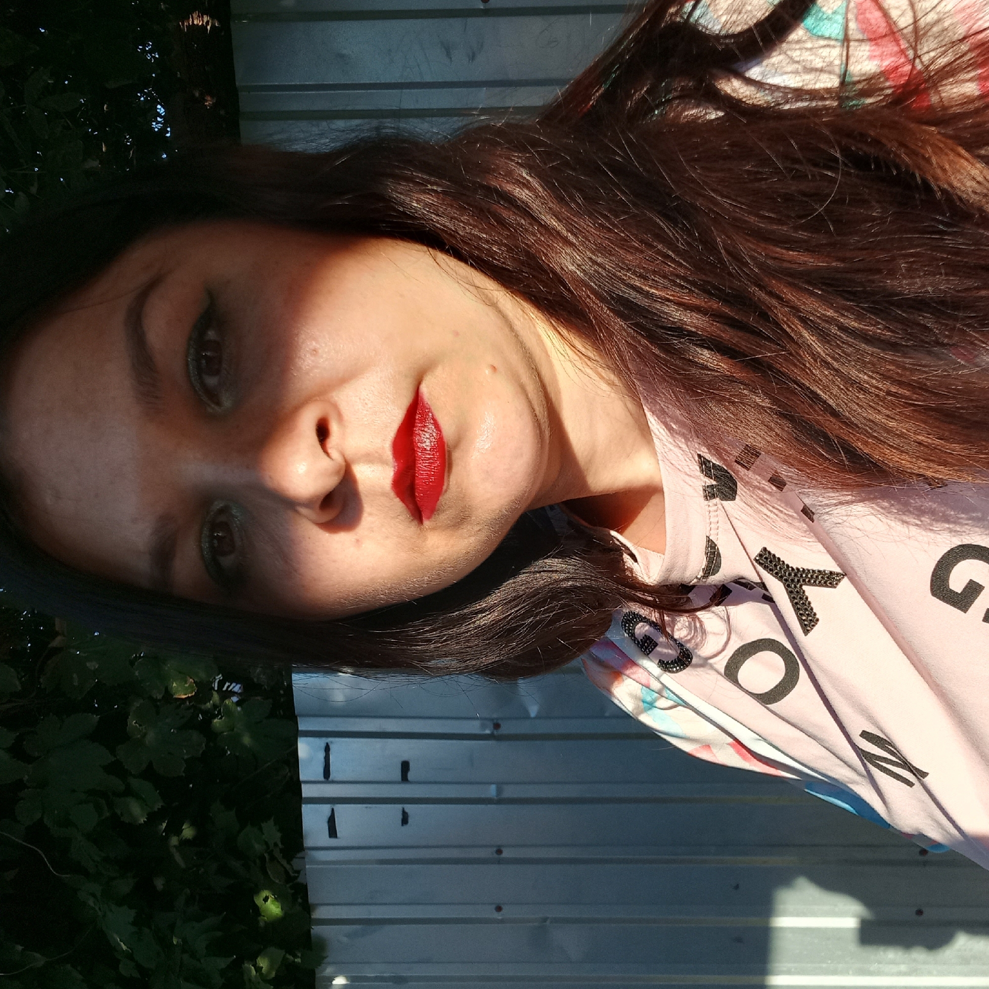 In Kropyvnytskyi, Ukraine, a woman with dark brown hair and makeup, including red lipstick and eyeliner, poses for a selfie outdoors. She is wearing a light pink t-shirt with black lettering and a colorful pattern. Behind her is a corrugated metal fence, and in the upper left corner, dark green foliage is visible. The sunlight casts a distinct shadow across her face, highlighting her features. The overall scene appears to be an informal, personal moment captured in an urban or suburban outdoor setting.