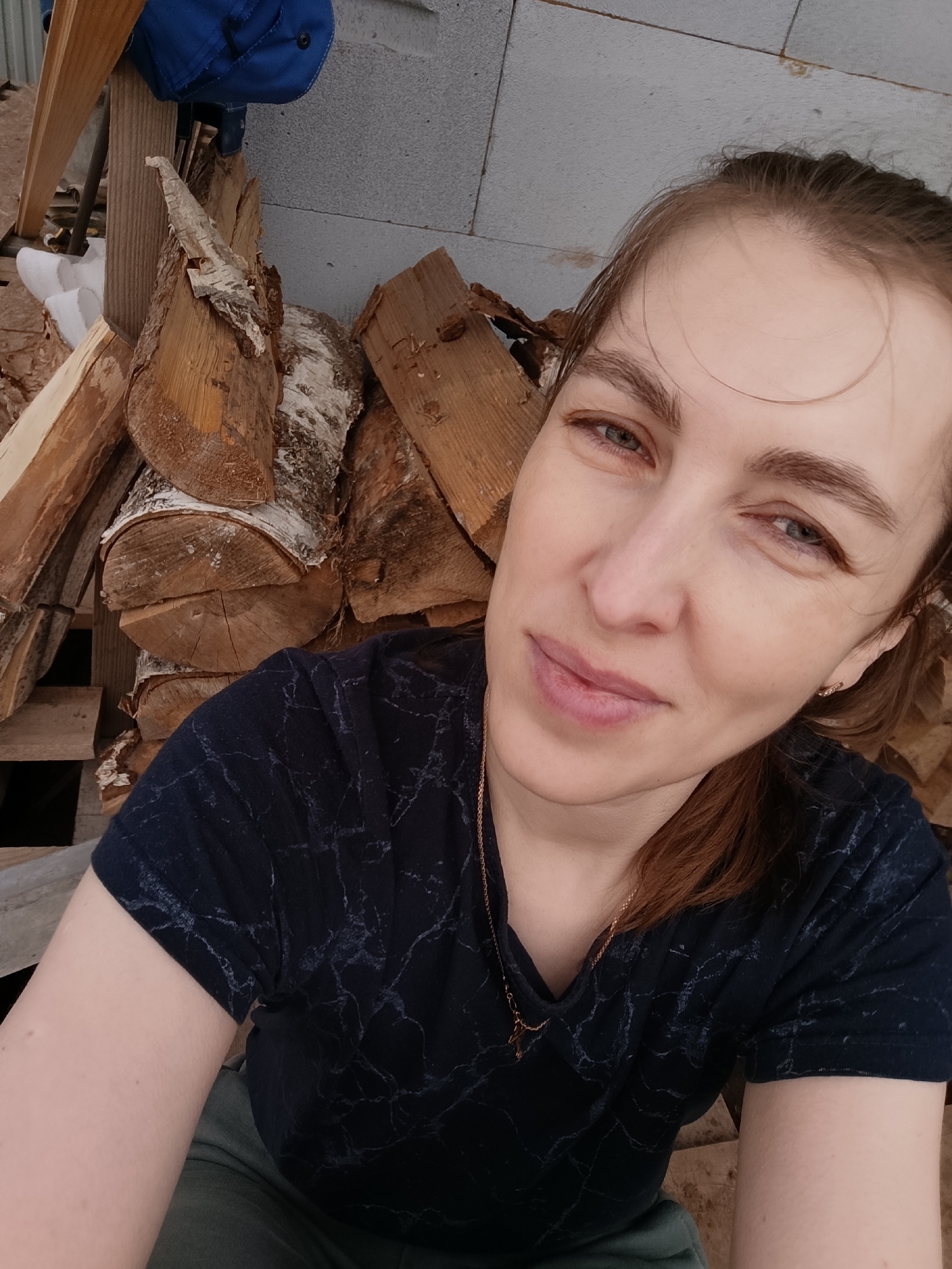 A woman is taking a selfie. She is positioned in front of a stack of firewood, and a concrete wall is visible behind the wood. A piece of blue clothing is visible at the top of the frame, partially obscured by the firewood. She is wearing a dark blue shirt with a marble-like pattern and a gold necklace with a pendant. The location is plausibly Chaikovsky, Russia due to visual cues in the environment.