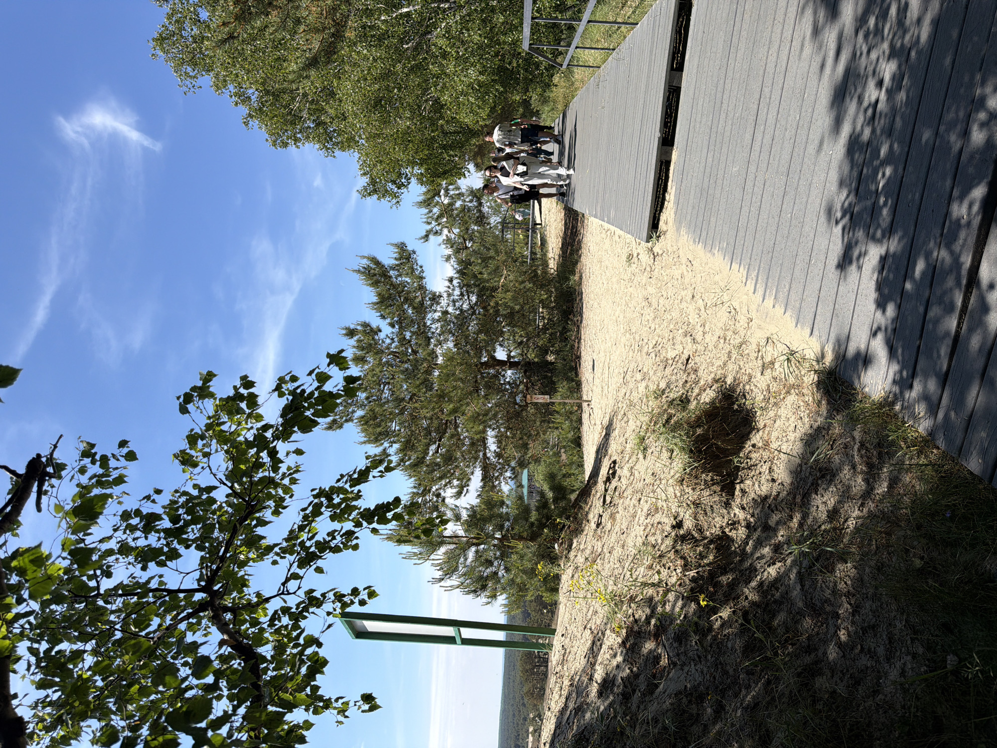 The image depicts a group of people walking on a gray wooden boardwalk that runs alongside a sandy dune area. The boardwalk is elevated and has metal railings on the right side. In the background, there are pine trees and a distant view of a forested area and possibly some buildings. The sky is blue with scattered white clouds. On the left side of the boardwalk, there is a sandy slope with sparse vegetation and shadows cast by trees overhead. A tall, green signpost stands on the sandy dune. The location is identified as Morskoe, Russia.