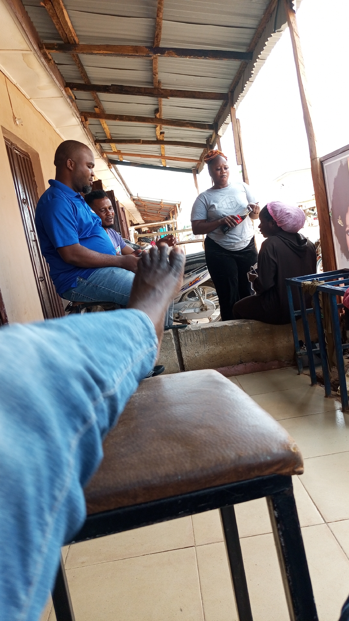 The image depicts an outdoor scene, likely in Sabon Tasha, Nigeria, based on the context. Four people are visible under a corrugated metal roof structure. A man in a blue shirt sits to the left, next to another man in a purple shirt. A woman in a grey shirt stands holding a Coca-Cola bottle and appears to be interacting with another woman wearing a pink head covering and a brown hoodie, who is seated on a low concrete structure. A motorcycle is parked behind them. A metal frame and a portrait are on the right side of the image. The foreground includes a leg in a blue jean resting on a worn-looking brown leather chair. The floor is covered with beige tiles.