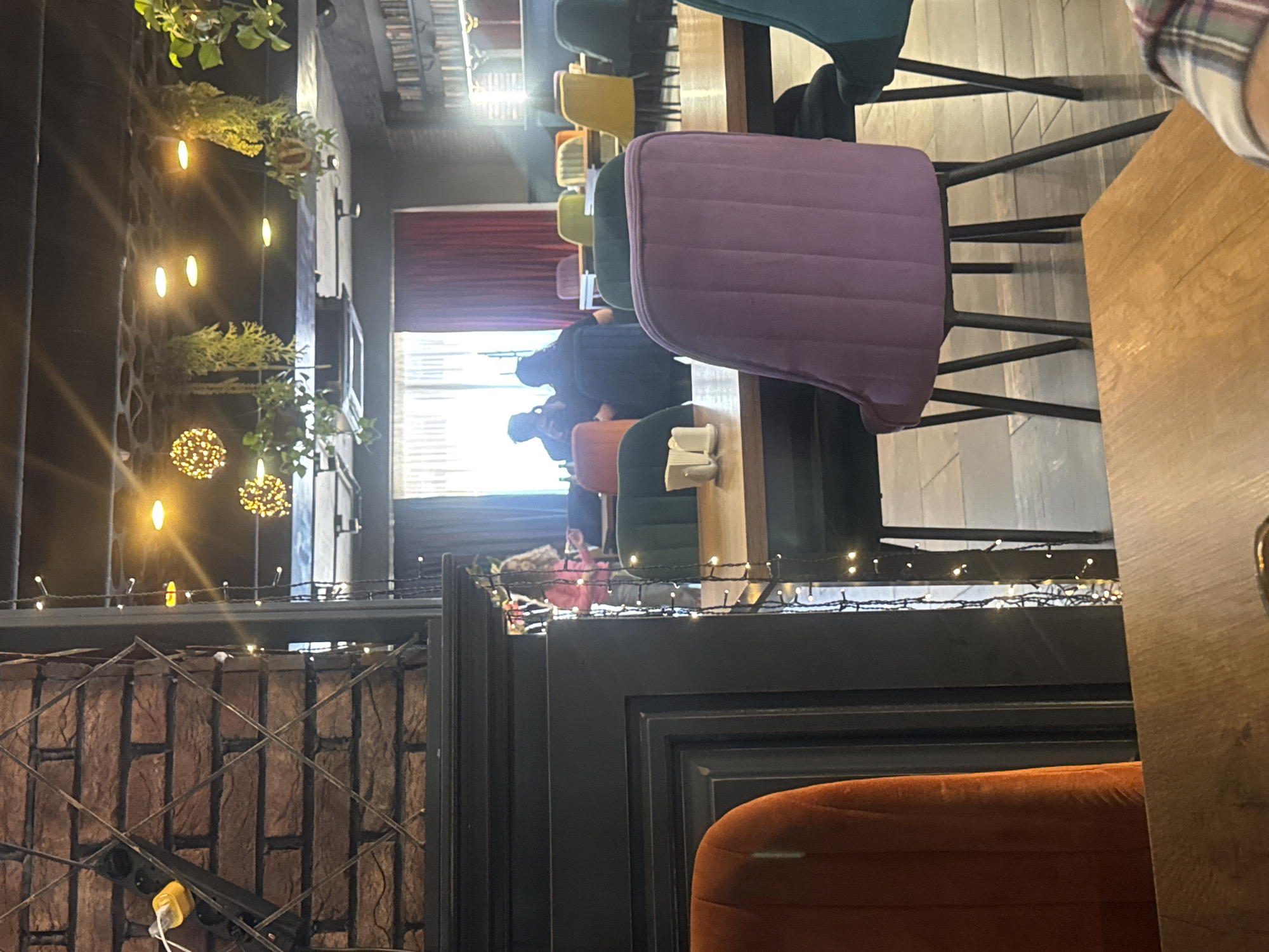 The image captures an interior scene in a restaurant or cafe, possibly located in Tbilisi, Georgia. The primary focus is on the seating arrangement, featuring a variety of chairs in different colors (purple, green, orange, turquoise, yellow) arranged around tables. People are seated at some of these tables, engaged in conversation or dining.
Architectural elements include brickwork, exposed metal structures, and decorative lighting (string lights and hanging lights with foliage). A curtain is visible in the background, suggesting a separation of spaces. The lighting creates a warm and inviting atmosphere.