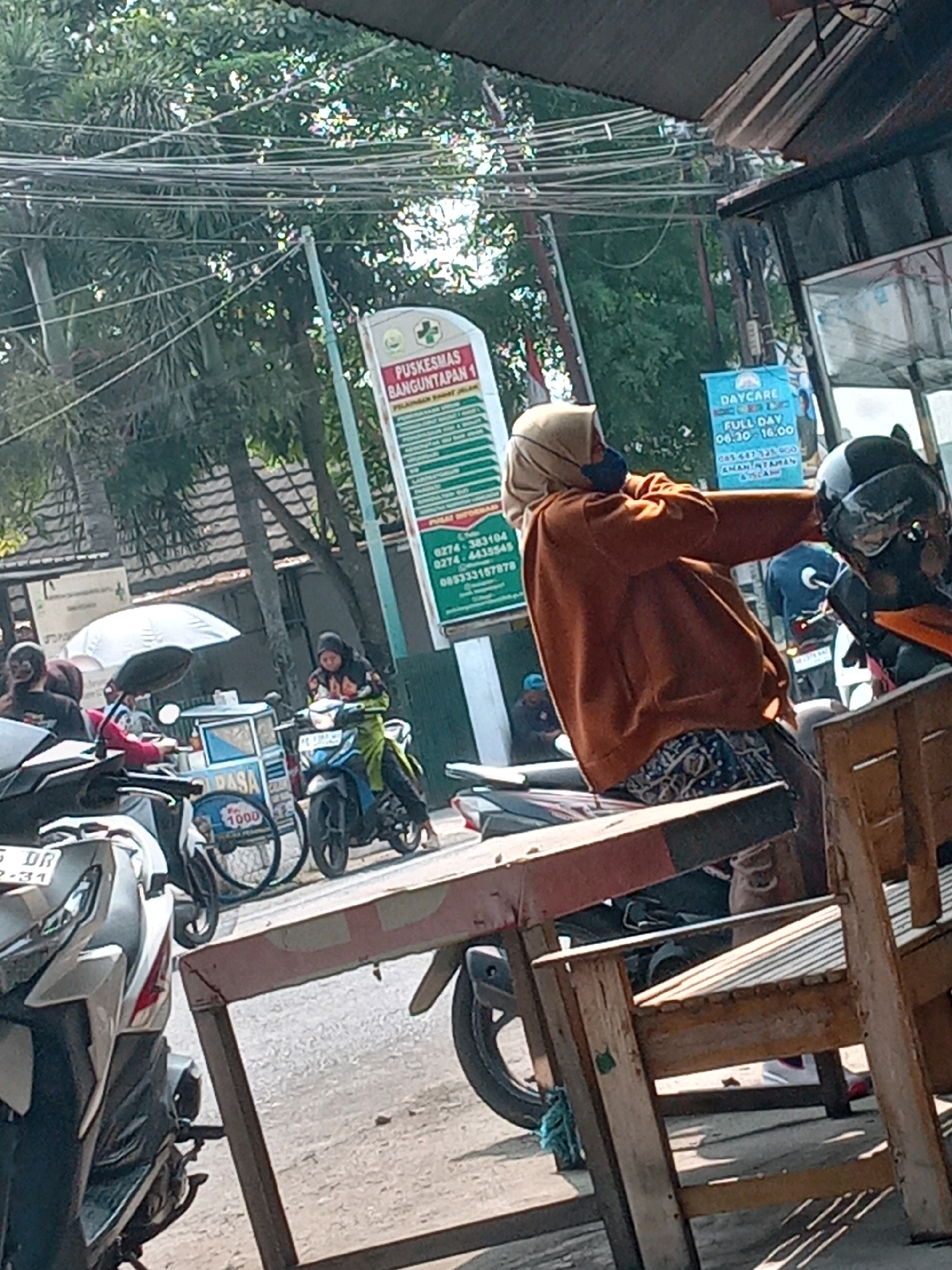 In Banguntapan, Indonesia, a street scene unfolds under bright sunlight. In the foreground, a person wearing a tan hijab and an orange sweatshirt is seated on a motorcycle, holding a helmet. The motorcycle has a black and white helmet on its seat. To the left, another person in a black shirt is on a motorcycle, and a small cart with the word "RASA" and "1000" is visible. In the background, a sign for "PUSKESMAS BANGUNTAPAN 1" (Health Center Banguntapan 1) is displayed along with another sign for "DAYCARE." Trees and power lines are overhead, and the ground appears to be paved. Other motorcycles and people are present in the scene, indicating a busy public area.