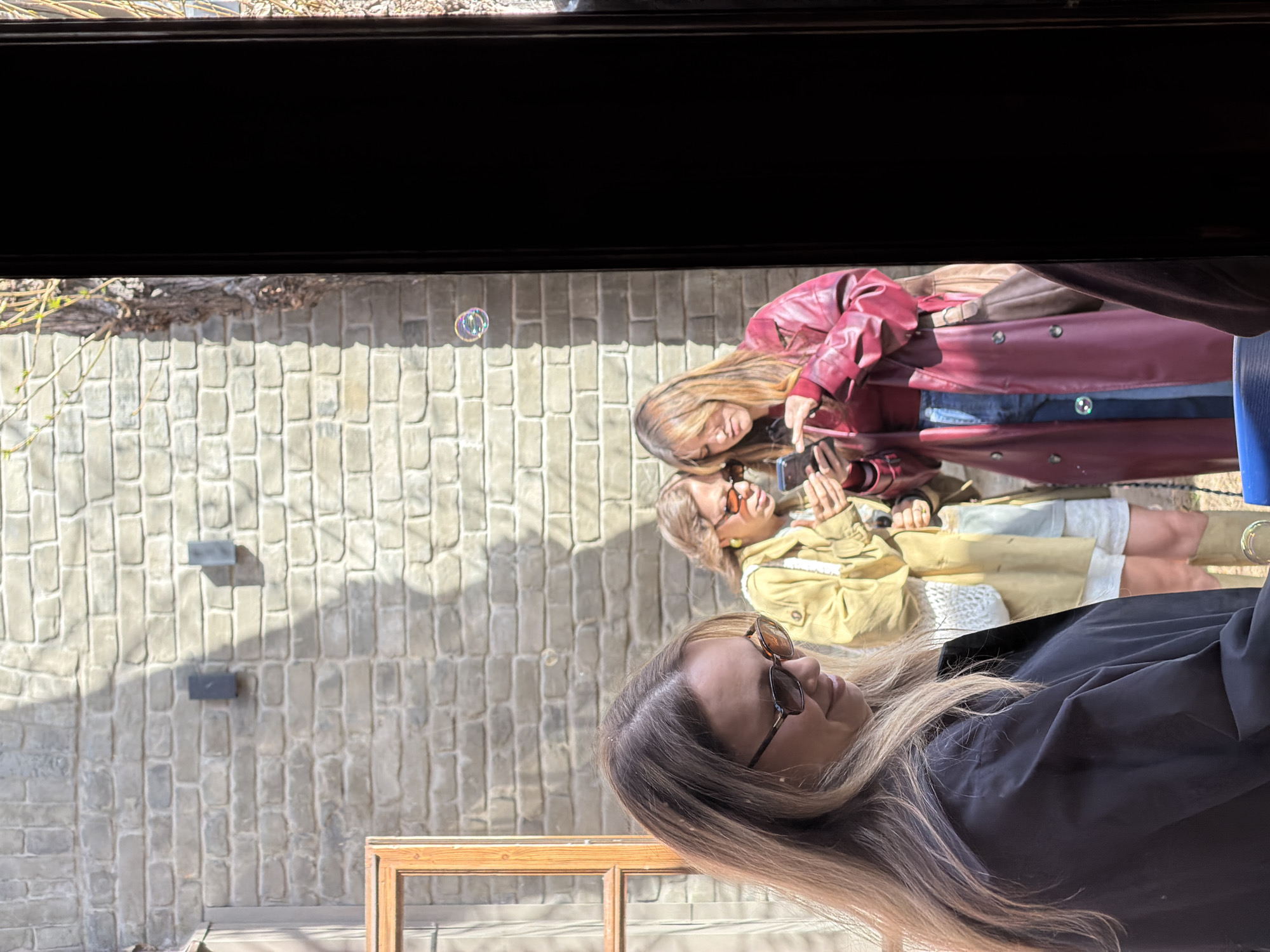 Three women are visible from behind a window frame in an outdoor setting. One woman closest to the camera is wearing a dark jacket and glasses. Two other women are taking photos or videos with a mobile phone; one woman is wearing a red jacket and the other a yellow jacket. A soap bubble is visible near a brick wall, which suggests the women are engaged in a playful activity. The exact location in Moscow, Russia, cannot be determined from the image alone.