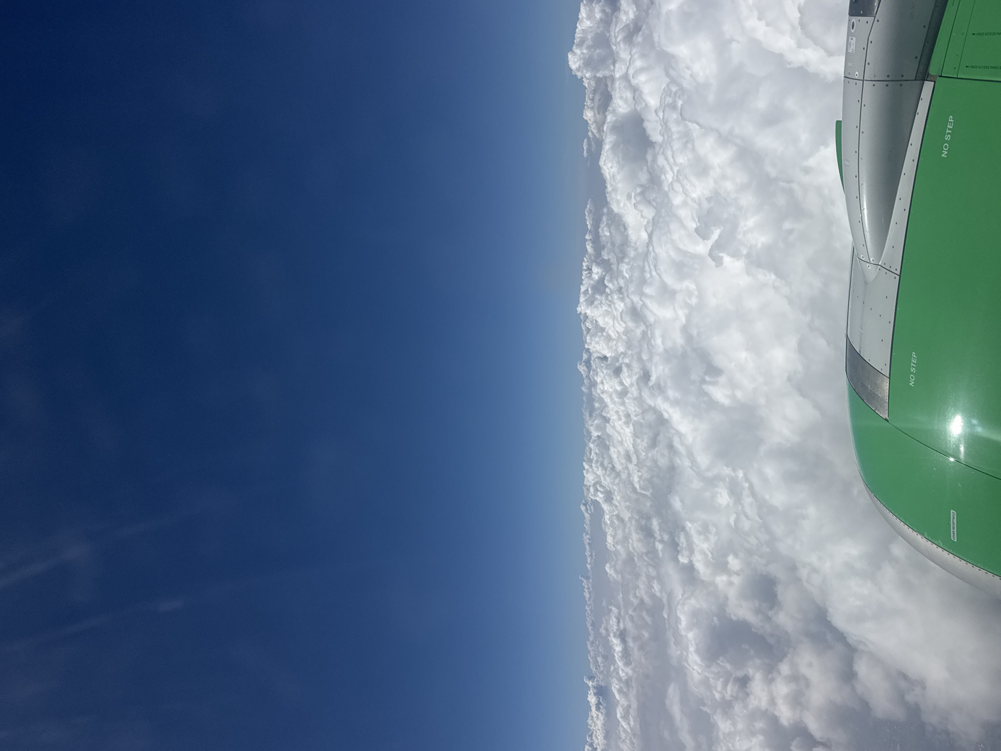 A high-angle view from an aircraft reveals a clear blue sky on the left, contrasting with dense white clouds on the right. The wing of the aircraft, painted green and gray, occupies the upper-right portion of the frame, featuring "NO STEP" markings. The absence of landmarks prevents determining if the location is near Epazoyucan, Mexico. There are no people or visible actions in the image.