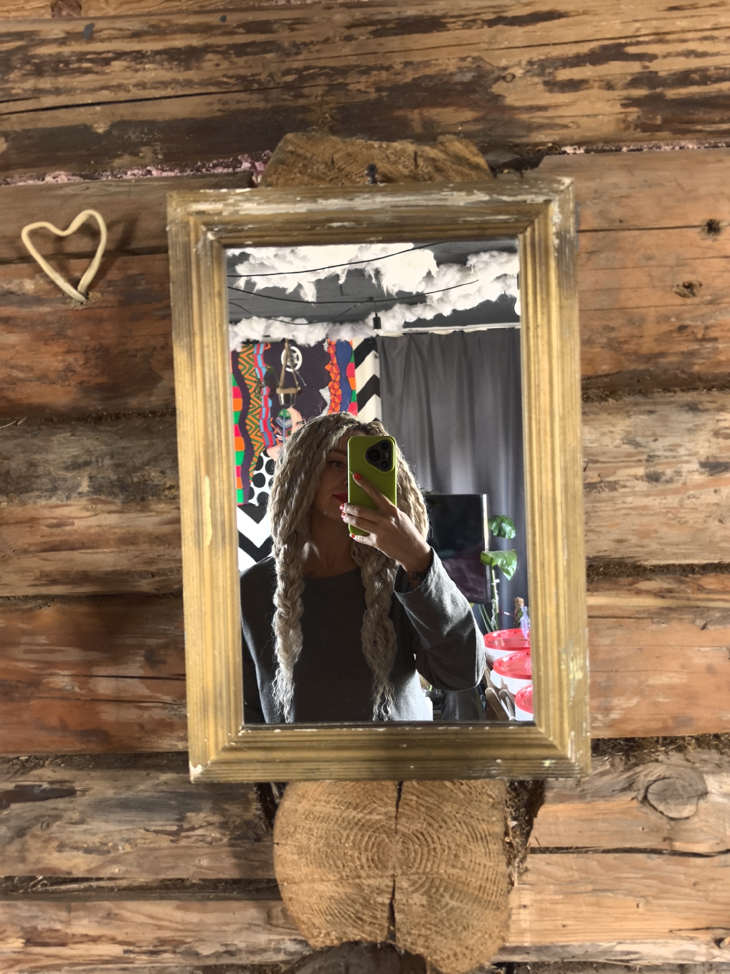 The image features a mirror selfie of a person with long, light-colored dreadlocks or braids, who is holding a bright green phone that partially obscures their face. They are wearing a dark grey long-sleeved top. The mirror itself has a distressed gold-colored frame and is mounted on a rustic wooden log wall, suggestive of a cabin or a traditional wooden structure. Below the mirror, the cross-section of a large wooden log supports it, while to the left, a heart-shaped object made of light-colored material is attached to the wall.
The reflection in the mirror reveals an interior space, likely a room within a location like Noviy, Russia. The ceiling in the reflection is decorated with white, fluffy material, possibly artificial snow or insulation, from which black wires hang. Dark grey curtains are visible, along with a flat-screen television on a stand and a plant with large green leaves. Further into the reflected background, a vibrant fabric tapestry or artwork featuring eclectic patterns like chevrons, polka dots, and stripes is displayed. Red and white plastic containers are also visible in the lower right of the reflection. The overall setting combines a rustic exterior with a personalized and eclectically decorated indoor environment. No text is visible in the image.