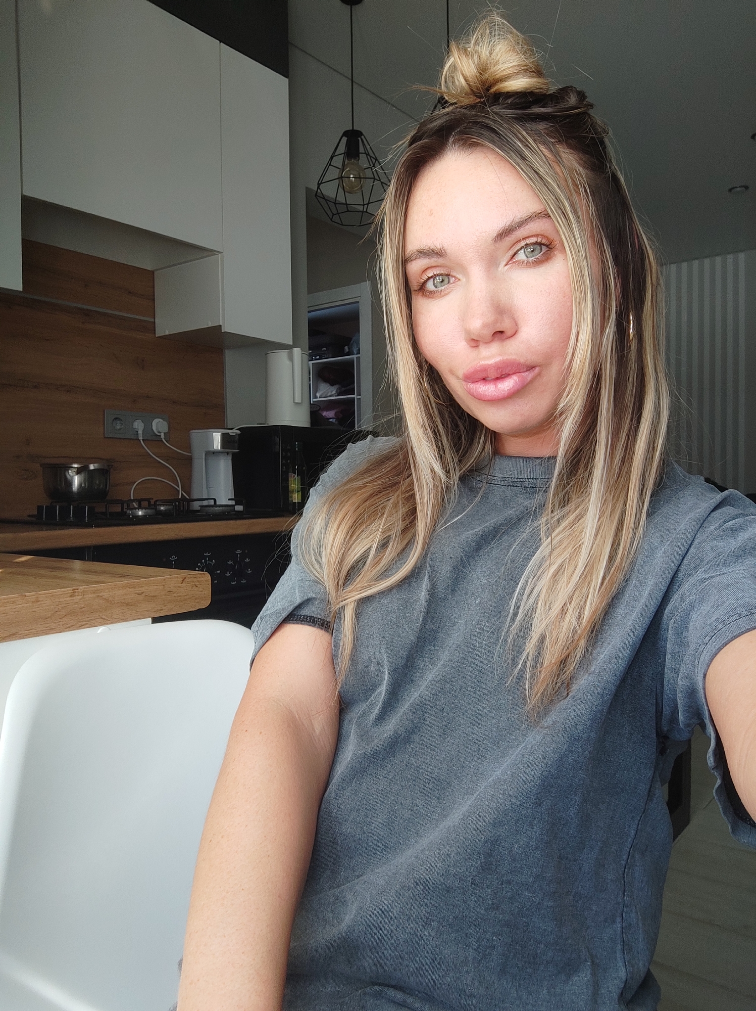 The image depicts a woman taking a selfie in what appears to be a modern residential kitchen or kitchenette located in Votkinsk, Russia. The woman, estimated to be in her mid-20s to mid-30s, has fair skin, light green eyes, and full lips with a light pink gloss. Her long hair is multi-toned with blonde and brown highlights, partially styled into a messy bun on top of her head while the rest falls over her shoulders. She wears a faded dark grey, short-sleeved t-shirt and a gold hoop earring is visible on her right ear. She is looking directly at the camera with a slight, neutral smile.
The background features a contemporary kitchen setup. To the viewer's left, a light wood-grain countertop holds a stainless steel pot on a black gas stovetop. Above the counter, a grey wall outlet with two sockets is visible, with power cords connecting to a small white coffee maker and a black appliance, possibly a microwave. A white electric kettle stands beside these items. White upper cabinets with a dark grey or black top trim are mounted above the counter. To the viewer's right, a wall with subtle light grey vertical stripes is visible. A dark, geometric metal cage pendant light fixture with an exposed filament bulb hangs from the ceiling. A partially visible doorway or opening in the far right background reveals shelving with folded materials. A portion of a white chair's backrest is present in the lower left foreground. The scene is brightly lit, suggesting natural light from an unseen window or strong artificial illumination.