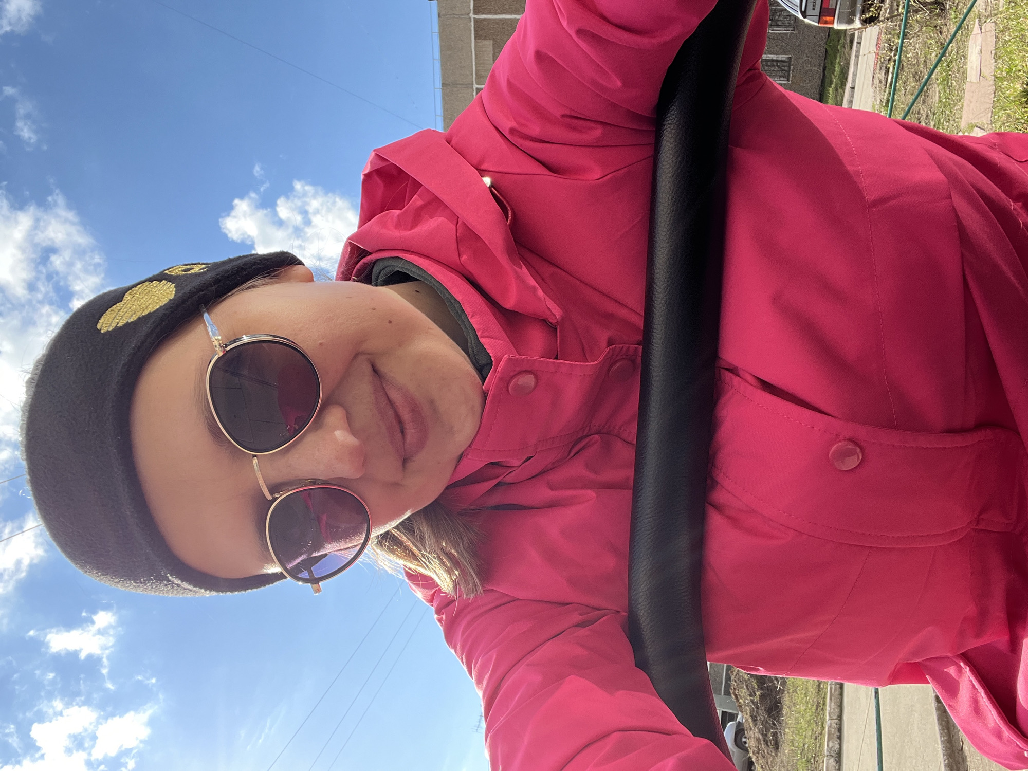 A low-angle selfie captures a person looking directly at the camera under a bright blue sky with scattered white clouds, suggesting sunny daytime weather. The subject, likely female, is wearing a vibrant hot pink hooded jacket, a black beanie with distinctive gold embroidered patterns, and round, dark-lensed sunglasses with gold-colored frames. A slight smile is visible, conveying a pleasant expression.
A prominent black bar or handle runs vertically along the right side of the frame, partially obscuring the person's torso and suggesting they might be seated in a vehicle or holding onto a railing. In the background, beyond the subject and the black bar, glimpses of an urban or suburban environment are visible. This includes parts of a building (possibly brick or concrete), some dry grass, and a partially visible car, indicating an outdoor setting in Chaikovsky, Russia. There is no visible text in the image.