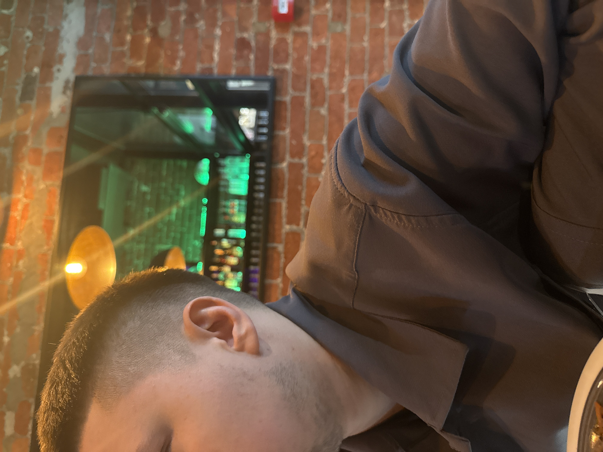 A close-up indoor shot shows a man with a close-cropped haircut, facing away from the camera. His right ear is visible. He wears a gray collared shirt or jacket. In the background is a brick wall with a red emergency light fixture near the top center. Behind him is a glass-enclosed structure with green lighting. The environment suggests an indoor location, possibly a restaurant or bar. There is no definitive information to identify this as being in Moscow, Russia.