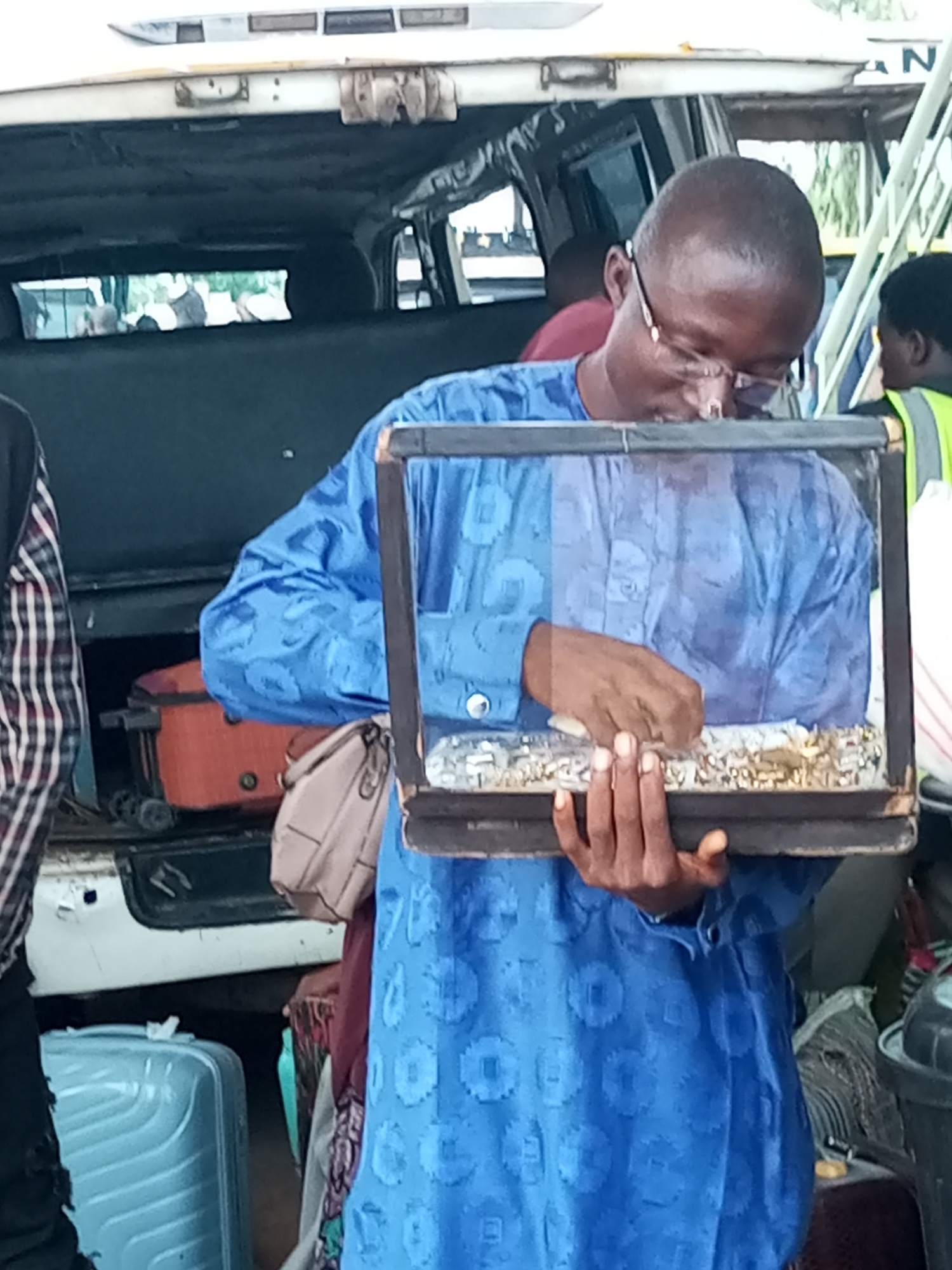 In Gombe, Nigeria, a man wearing a blue patterned tunic and glasses is holding a glass-encased object containing what appears to be a collection of small, shiny items, possibly seeds or beads. He is looking down at the object, with his hands near the contents. The scene takes place outdoors, with the back of a vehicle, possibly a bus or van, visible behind him. Luggage, including a blue suitcase and an orange travel case with wheels, is piled in the background. Another person, wearing a green vest, is visible to the right. The lighting suggests it is daytime. The overall impression is one of preparation for travel or a transaction involving the items in the case.