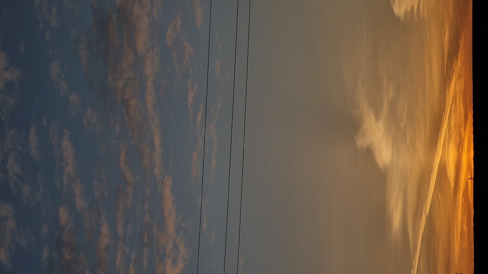 The image depicts a sunset over the city of Vysokoe, Russia. The sky is a gradient of colors, starting with a deep blue at the top, transitioning to a soft gray, and culminating in vibrant oranges and yellows near the horizon. Scattered across the upper sky are small, wispy clouds tinged with pink. Three dark, thin power lines stretch horizontally across the middle section of the image, dividing the sky into distinct zones. Towards the bottom, a dark silhouette of the landscape is visible, obscuring details of the ground and any structures. A contrail from an aircraft is faintly visible in the lower part of the sky, adding a diagonal streak to the scene. The overall impression is one of a serene, twilight atmosphere.