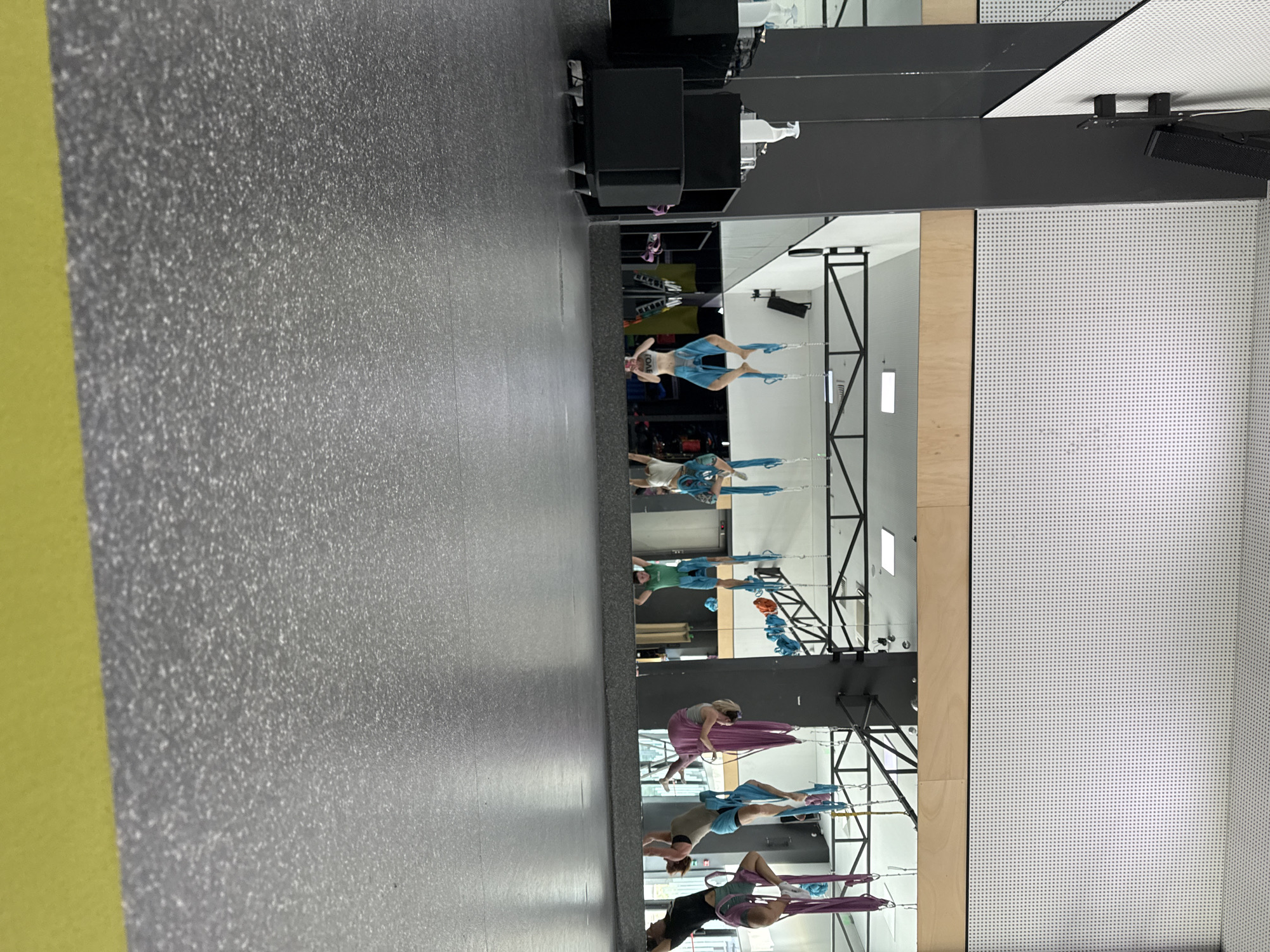 In an indoor gymnasium with a grey speckled floor and a yellow border along the bottom, several individuals are engaged in aerial yoga. The gymnasium features a large mirrored wall reflecting the activity. Above, a metal frame supports multiple fabric hammocks in various colors, including blue, purple, and pink. One person, positioned in the center of the reflection, is upside down in a blue hammock, wearing blue shorts and a dark sports bra. To their right, another person is similarly suspended in a purple hammock, wearing grey leggings and a grey top. Further to the right, a third person is in a blue hammock, wearing a grey sports bra and grey shorts. On the left side of the reflection, another individual is suspended upside down in a blue hammock, wearing blue shorts and a light-colored top. A fourth person is in a green hammock, wearing a green tank top. The scene is brightly lit, suggesting an active or training environment.