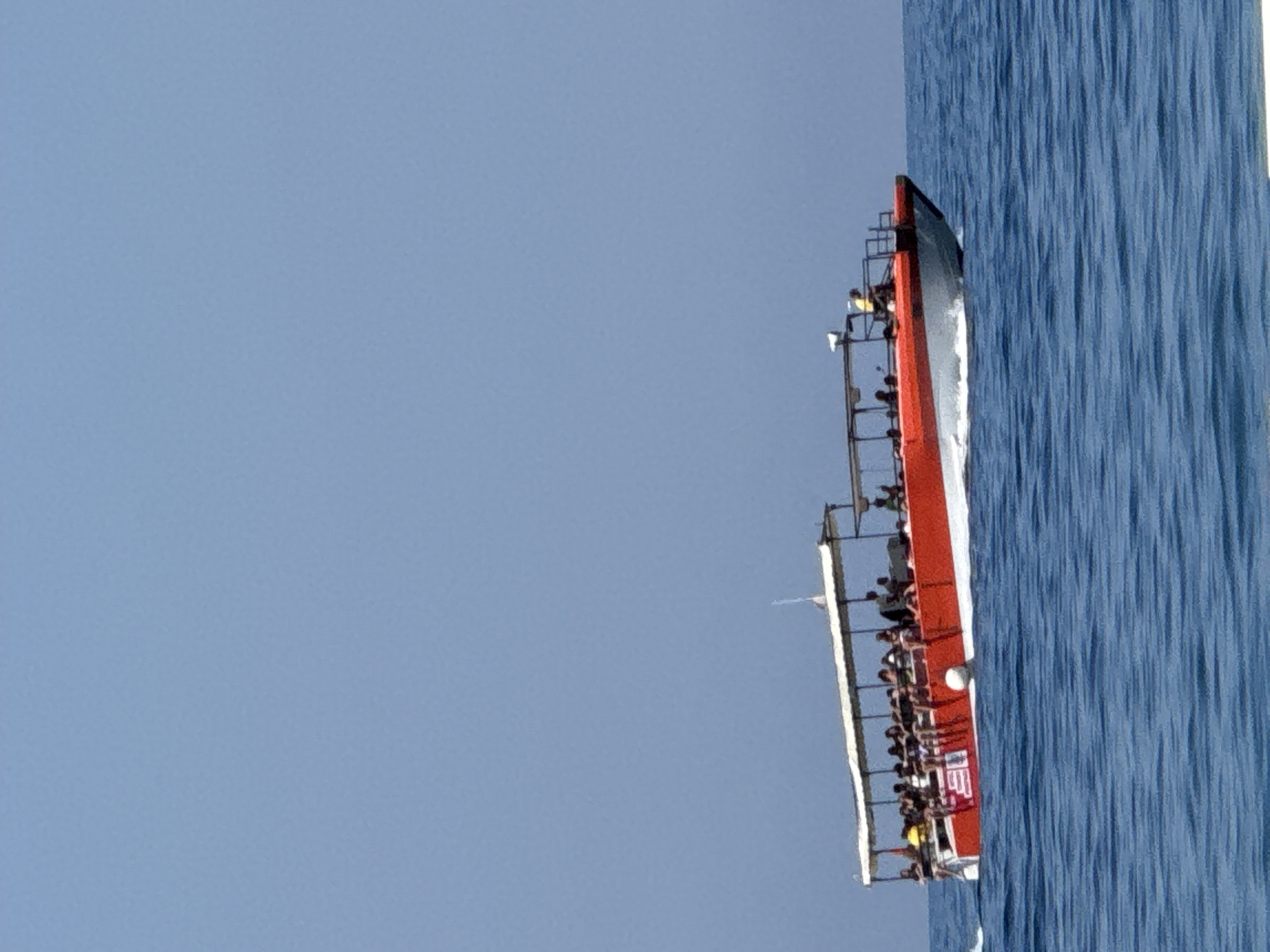 In this image, a red and white vessel with a superstructure is sailing on a calm, blue sea. The vessel is long and narrow, with multiple decks visible. A number of people are standing on the decks, some appearing to be passengers or crew. The vessel has a distinctive red stripe along its side and a white superstructure with railings. There is also some scaffolding or a tower-like structure on the vessel, with lights attached. The water is a deep blue with a textured surface, suggesting gentle waves. The sky above is clear and pale blue, indicating a bright, possibly sunny day. The perspective is from slightly above and to the side of the vessel, looking down its length. The image is oriented vertically, with the vessel running from the top right to the bottom left. A portion of what appears to be a flag or banner with the letters "US" is visible on the side of the vessel. The scene is tranquil and suggests a leisurely journey on the water.