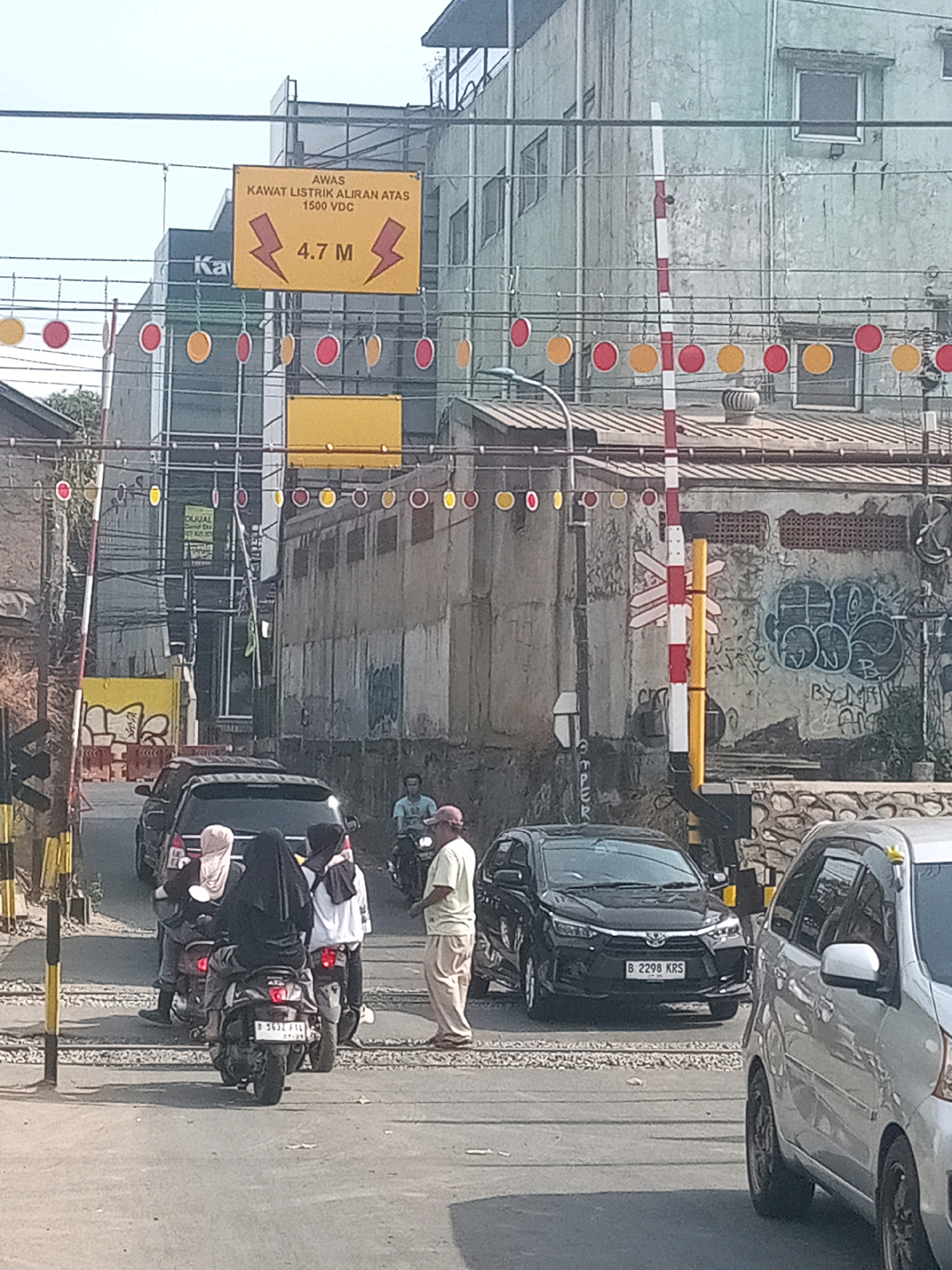 The image depicts a street scene in Bekasi, Indonesia, likely near a railway crossing. In the foreground, a silver van is parked, with its front right wheel visible. To the left, a black SUV is positioned on the street. Behind the SUV, three people are on a motorcycle; two are passengers wearing headscarves, and one is the driver. A man on a motorcycle, wearing a light blue shirt and a pink cap, is to the right of the black SUV.
Dominating the mid-ground are several prominent signs and structures. A yellow warning sign with red lightning bolts and the text "AWAS KAWAT LISTRIK ALIRAN ATAS 1500 VDC 4.7 M" indicates overhead high-voltage electrical wires with a clearance of 4.7 meters. This sign is mounted on a pole. Adjacent to it, a red and white striped pole, likely part of a railway crossing barrier, stands tall. Below the warning sign, a yellow rectangular sign is visible. A line of hanging red and yellow circular markers stretches across the scene, suggesting a festive or cautionary decoration.
The background reveals buildings characteristic of urban Indonesia, with some appearing weathered and others more modern. Graffiti is visible on the wall of a building in the mid-ground to the right. The overall environment suggests a busy urban street with evidence of industrial or electrical infrastructure. The lighting indicates it is daytime.