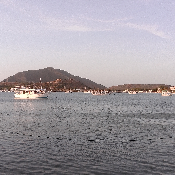 A seascape under a hazy sky. Several white boats are scattered across the water, with a larger one prominent in the foreground. Mountains form the backdrop, marked by some man-made structures. The water is relatively calm, with hints of ripples, and the horizon line is clear.