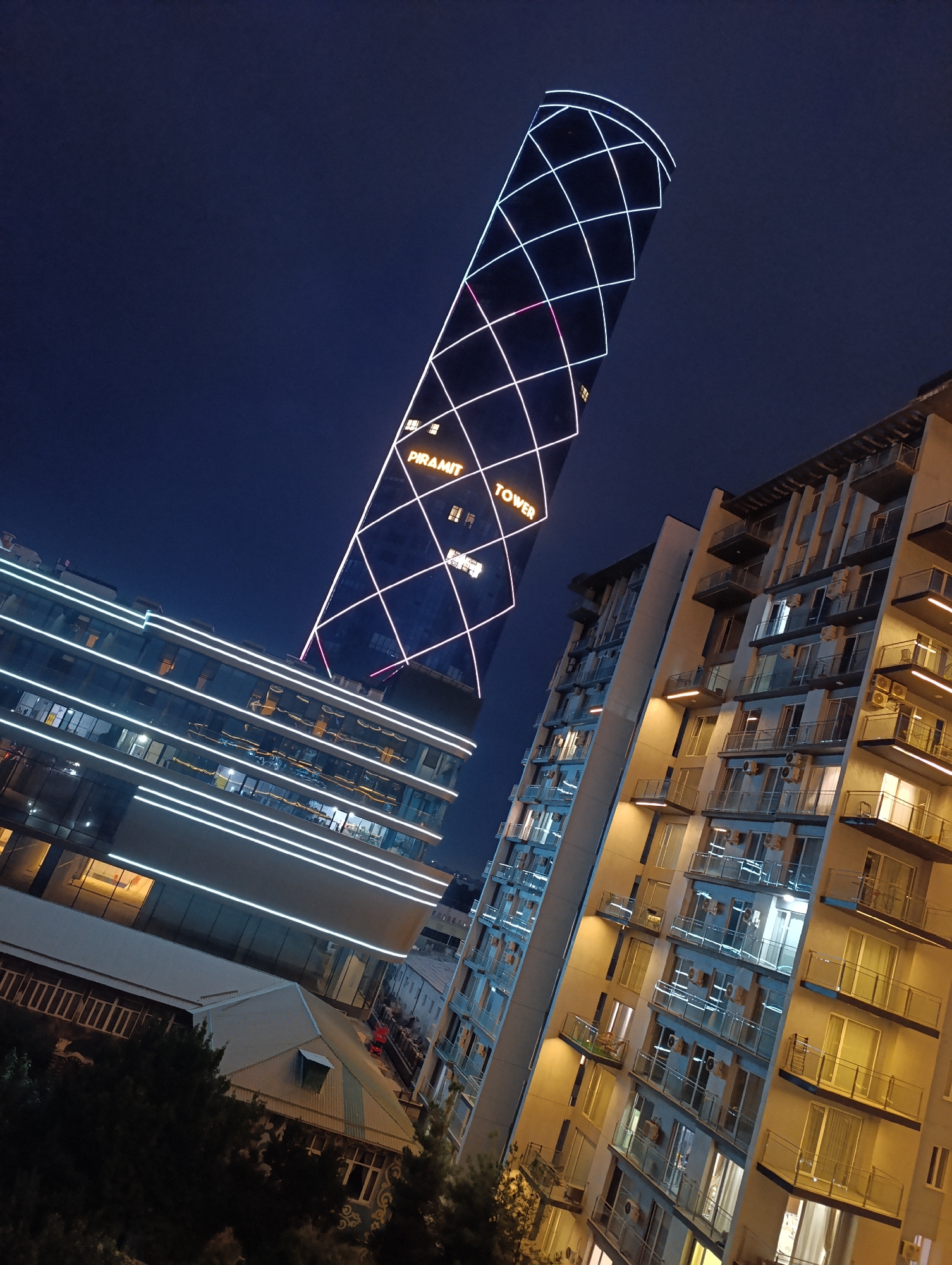 The image depicts a nighttime cityscape in Tashkent, Uzbekistan, featuring the Piramit Tower. The Piramit Tower, a distinctive skyscraper with a diagonal grid of white lights and illuminated text spelling "PIRAMIT TOWER", dominates the upper portion of the frame. To the left of the tower is a modern glass-facade building with horizontal white light strips. To the right, a residential building with multiple balconies, air conditioning units, and illuminated windows is visible. The sky is a deep blue, indicating it is night. Trees are present in the foreground, obscuring the lower parts of some structures. There are no people visible in the image. The scene is one of urban architecture illuminated at night.