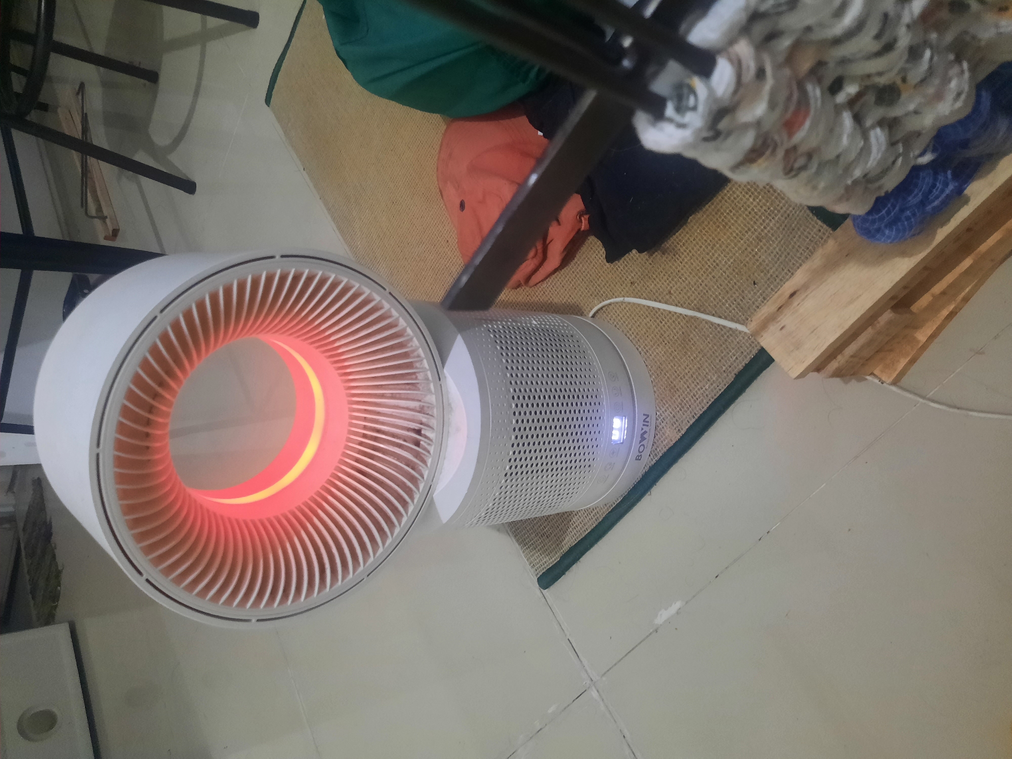 The image shows a white fan heater with a glowing orange inner ring, positioned on a woven mat on a tiled floor. The fan has a perforated base with a digital display showing "BOWIN". In the background, there are furniture legs, a green fabric, and orange and black clothing. To the right of the fan, there is a stack of wooden planks and a mop. The scene appears to be an indoor domestic setting, possibly a living room or bedroom, in Sidokepung, Indonesia. There are no people visible in the image. The lighting suggests it is either evening or the room is dimly lit.