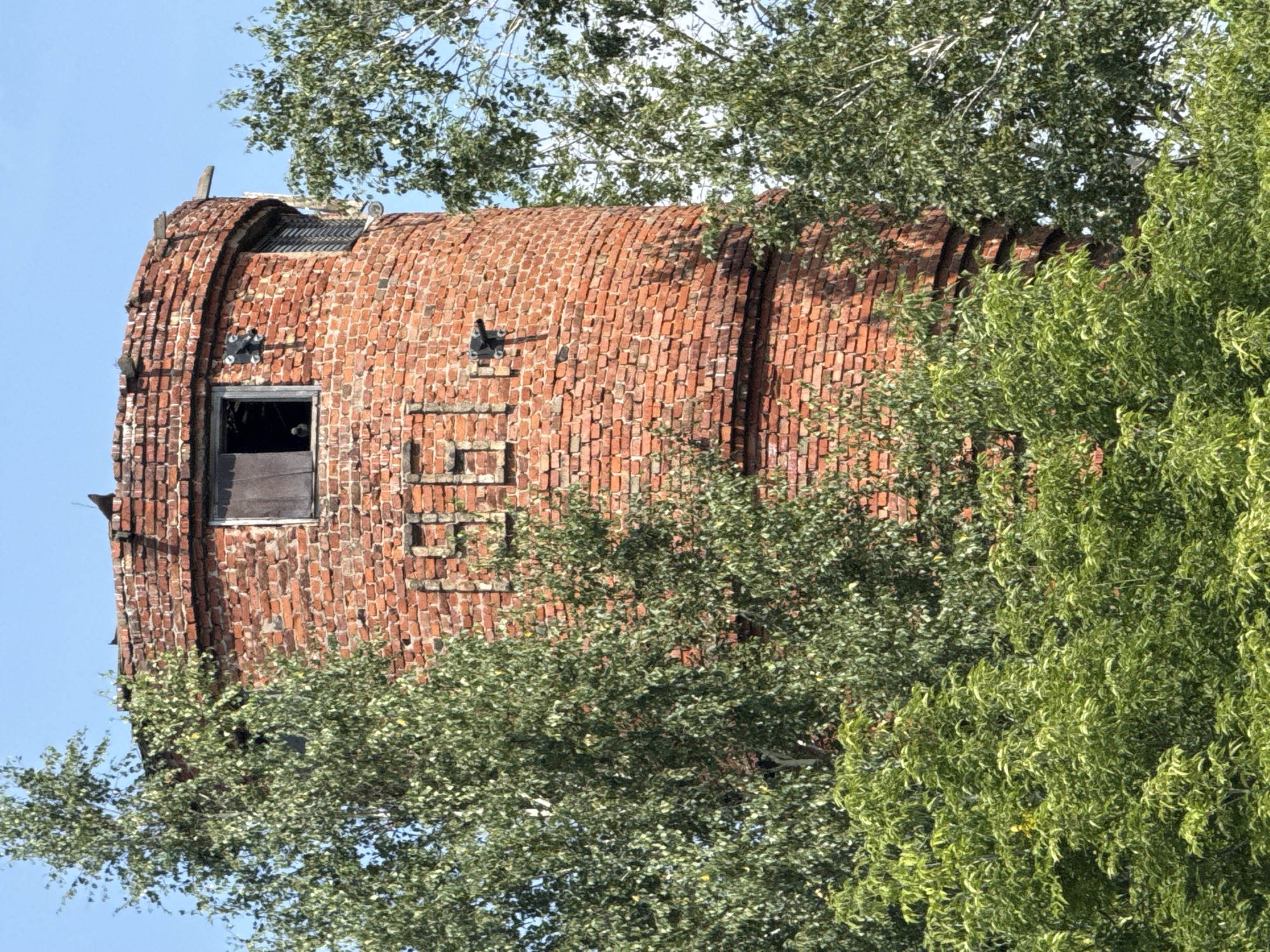 An old, red brick tower, possibly a water tower or part of an industrial structure, stands partially obscured by lush green foliage. The tower is cylindrical and shows signs of disrepair, with broken window frames and missing roof elements. Inlaid into the brickwork are the numbers "1961". Two black fixtures, possibly for lighting or cameras, are mounted on the exterior. The sky above is clear and blue. The scene is located in the city of Osa, Russia.