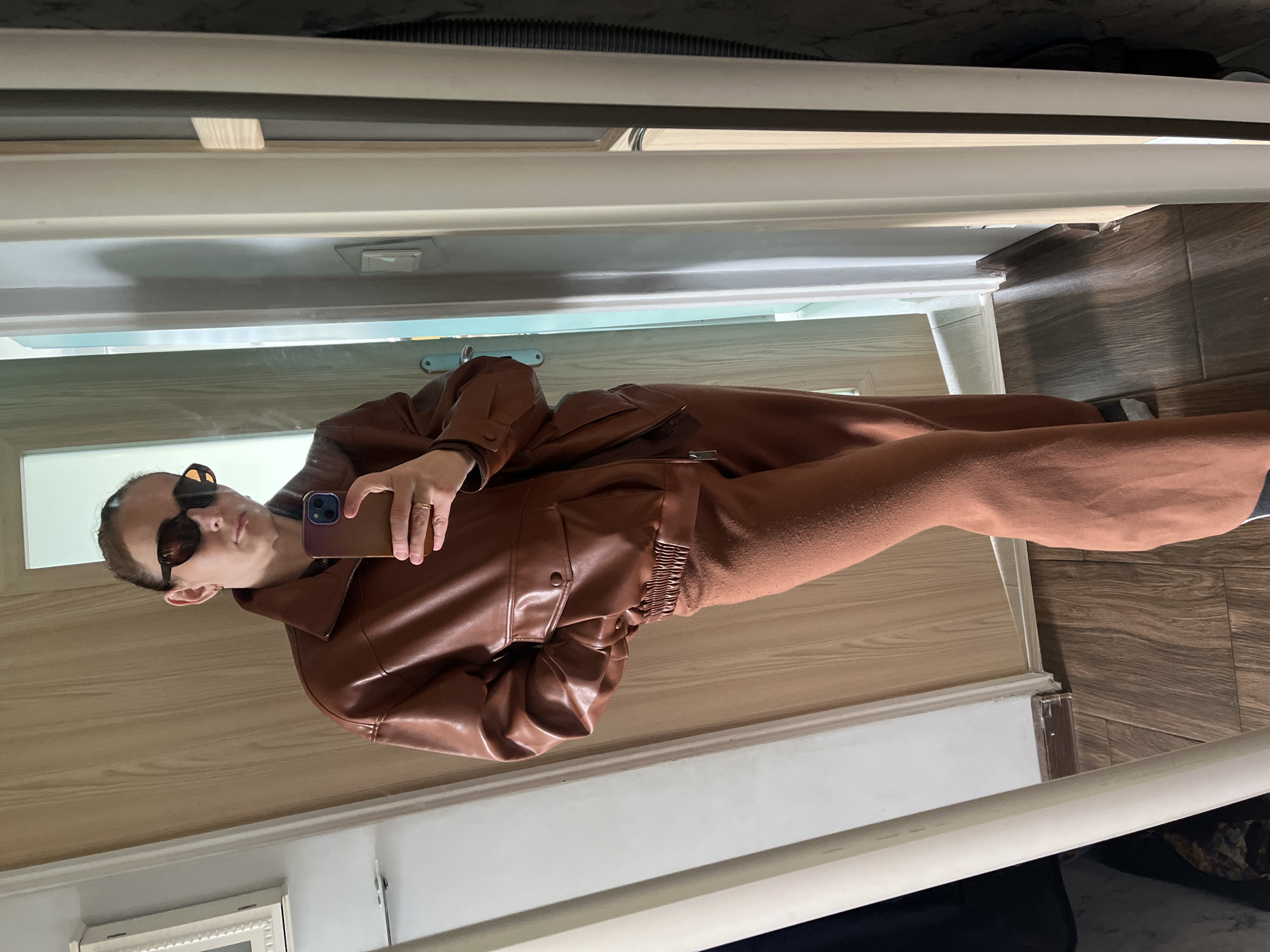 A person is visible in a mirror, taking a selfie. The individual wears sunglasses, a brown leather jacket, and what appears to be matching brown pants. The reflection shows the person holding a smartphone in their right hand. The background includes portions of a doorframe, wooden flooring, and what might be a wall or another surface with a white trim. The location is unknown.