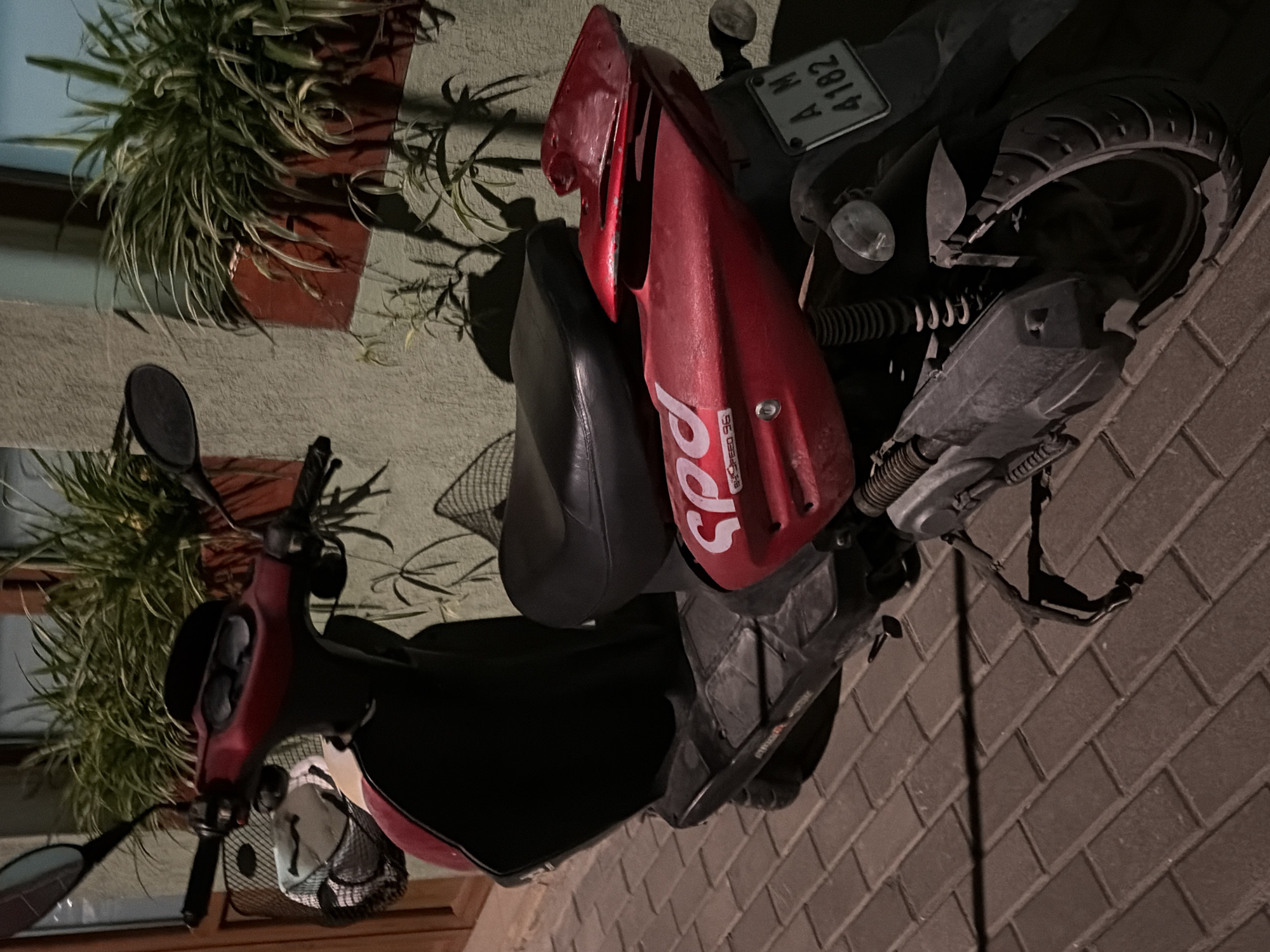 In the city of Obzor, Bulgaria, a red scooter is parked on a brick sidewalk. The scooter is facing left, with its front wheel angled slightly towards the right. The scooter has a black seat and a black front panel. On the right side of the scooter, a license plate with the letters "AM" and the numbers "4182" is visible. Behind the scooter, a light green wall is visible, with green plants in planters on the wall. A wooden door is visible on the left side of the image. The lighting suggests it is nighttime, with some artificial light illuminating the scene.