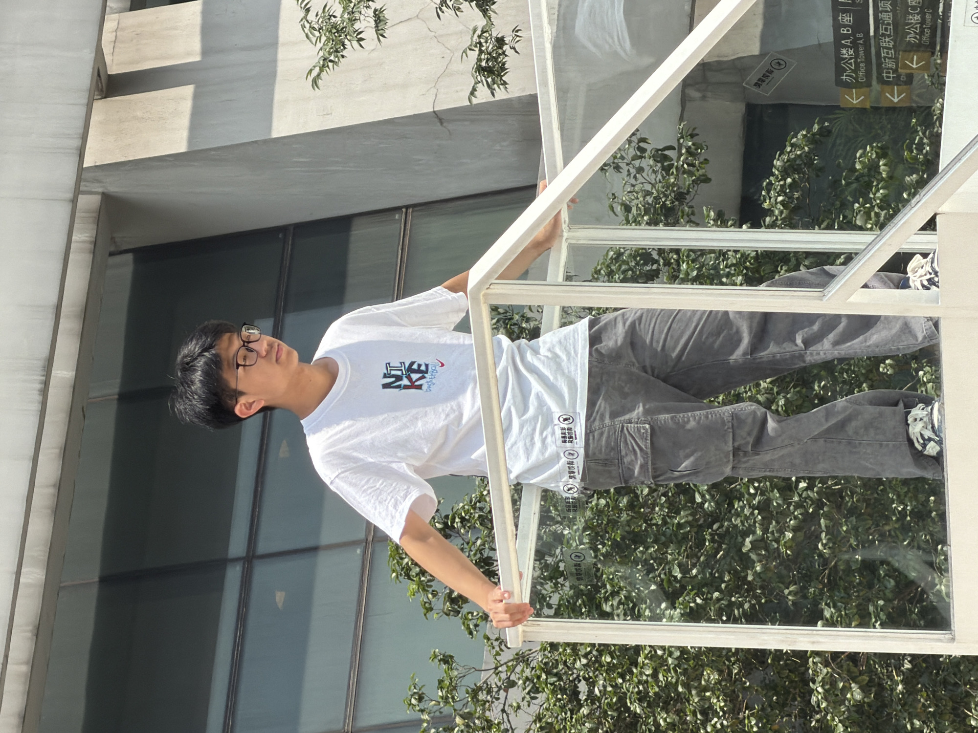In Yuzhong District, China, a young East Asian male wearing a white t-shirt with a graphic print and dark cargo pants stands on a white railing overlooking lush green foliage. He is wearing glasses and his hands rest on the railing. The background features a modern building with large glass windows and concrete architectural elements. Signs indicating "Office Tower A, B" and "Office Tower C" are visible in the distance, suggesting this is a commercial or office complex. The lighting indicates it is daytime, with bright sunlight casting shadows.