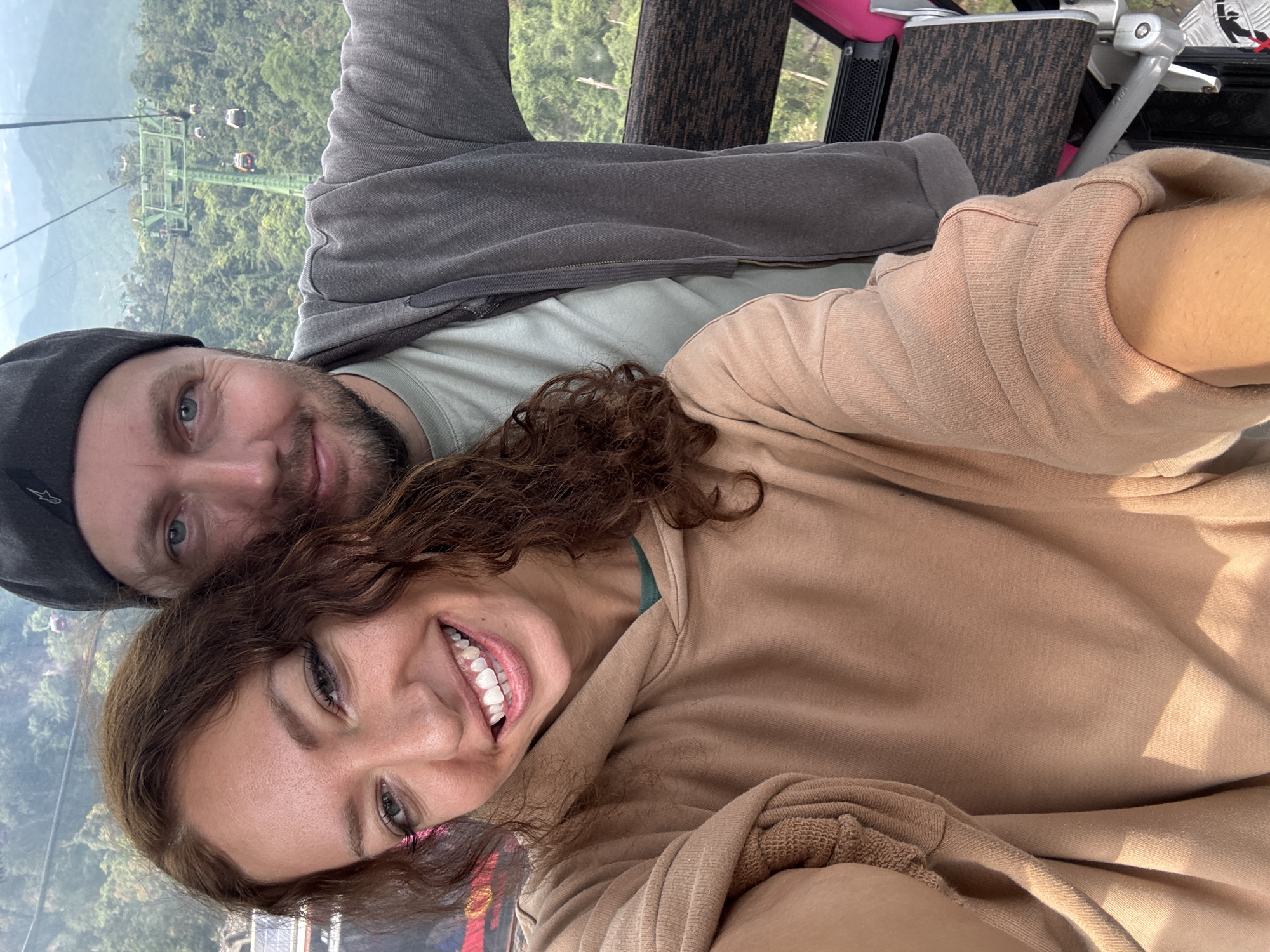The image captures a joyful selfie of a man and a woman inside a cable car, positioned above a vast, lush green landscape. The shot is taken from a slightly upward angle, with both individuals smiling directly at the camera.
The man, on the left side of the frame, has a light beard, blue eyes, and is wearing a dark grey beanie, a light green t-shirt, and a grey hooded jacket. He has a warm smile. The woman, on the right, has dark, curly hair, a bright, wide smile showing her white teeth, and appears to be wearing light makeup. She is dressed in a light brown hooded sweatshirt, with a glimpse of a light green garment visible underneath the collar.
Behind the couple, through the cable car window, a dense, verdant forest blankets rolling hills or mountains. A prominent cable car system with several towers and other cabins in the distance stretches across the landscape, suggesting a significant elevation. Parts of the cable car's interior, including a brown patterned seat, metallic structures, and a window frame, are visible around the subjects.
The activity taking place is a leisure ride on a cable car, likely a sightseeing experience. The bright lighting, clear visibility, and vibrant green foliage indicate a pleasant daytime. Both individuals exhibit happiness and contentment, reflecting positive emotions. Given the provided context of Đà Nẵng, Vietnam, this is likely a scene from a popular tourist destination like the Bà Nà Hills cable car. No discernible text is clearly visible, though the man's beanie has a small, indistinct logo.