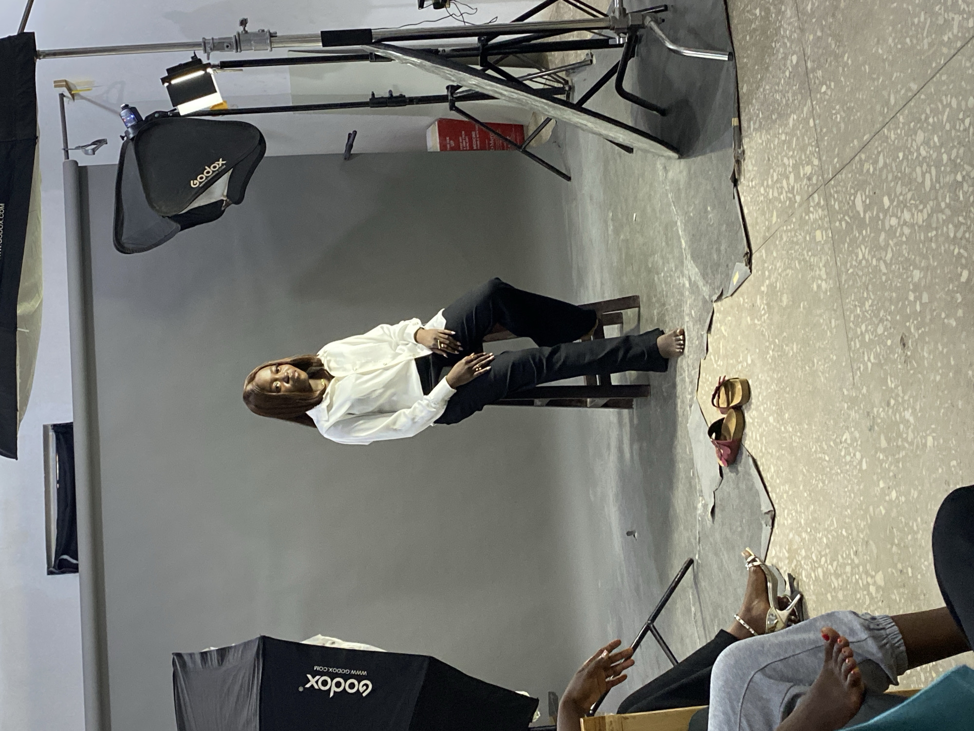 The image shows a studio setting, possibly in Jos, Nigeria, due to the provided information. A woman with brown hair is posing in a white blouse and black pants, seated on a wooden chair against a grey backdrop. Several studio lighting equipment pieces are visible, including two Godox softboxes on stands and a reflector. To the side, a pair of sandals and high heels are placed on the floor. Parts of other individuals (hands, feet, and legs) are visible, indicating they are likely observers or crew members in the studio. The floor is a textured grey and white. The background is a solid grey photography backdrop.