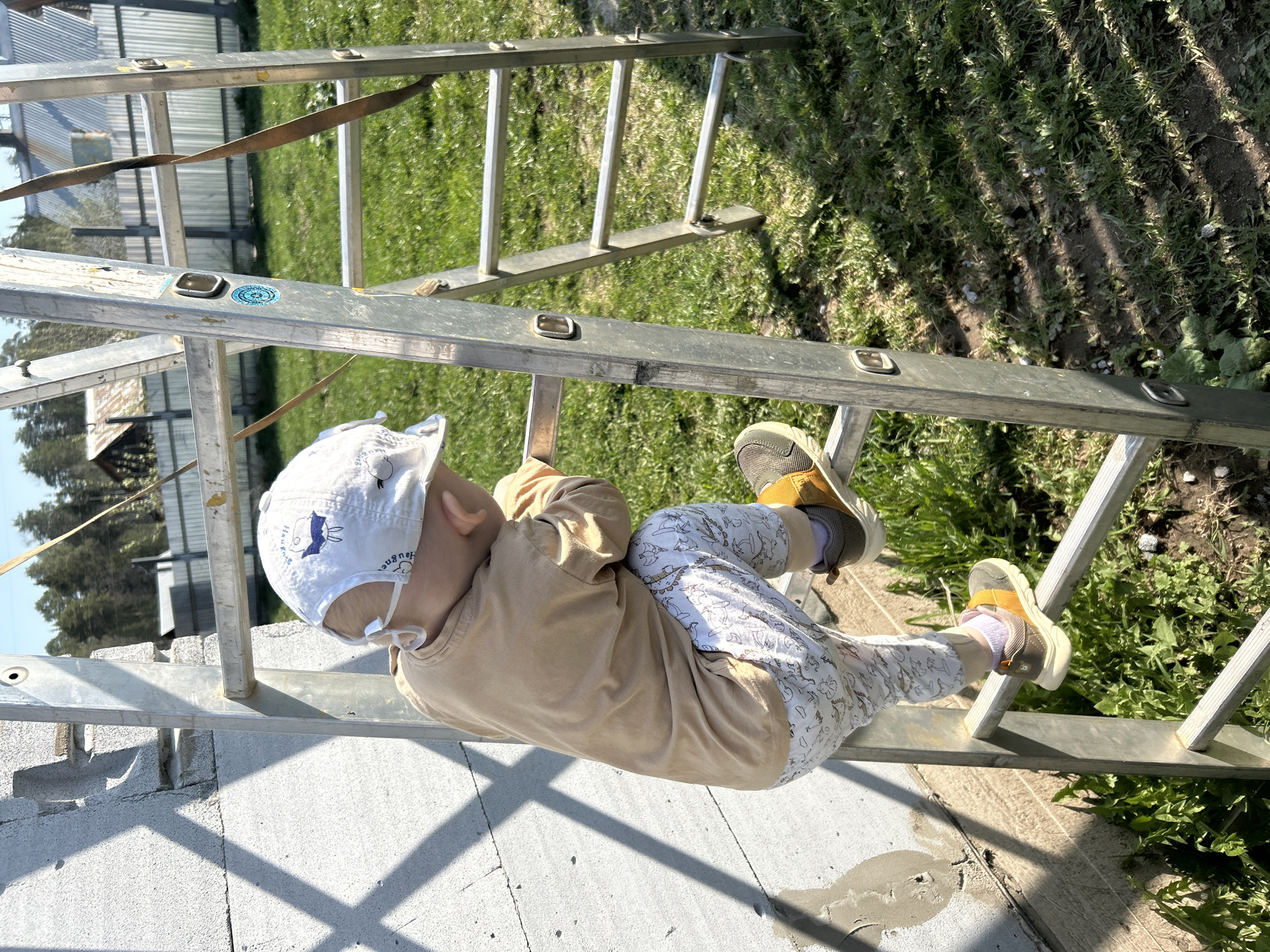 A child wearing a white cap, beige long-sleeve shirt, and patterned pants is precariously climbing an aluminum extension ladder. The ladder is leaning against a gray cinderblock wall, with grass and foliage visible in the background. The location is outside, possibly in Chaikovsky, Russia, given the origin of the image. The child's feet, clad in yellow-strapped sneakers, are planted on the ladder's rungs. The ladder extends upwards towards a grassy area and is secured by a strap at the top. The wall beneath the ladder shows signs of construction or renovation.