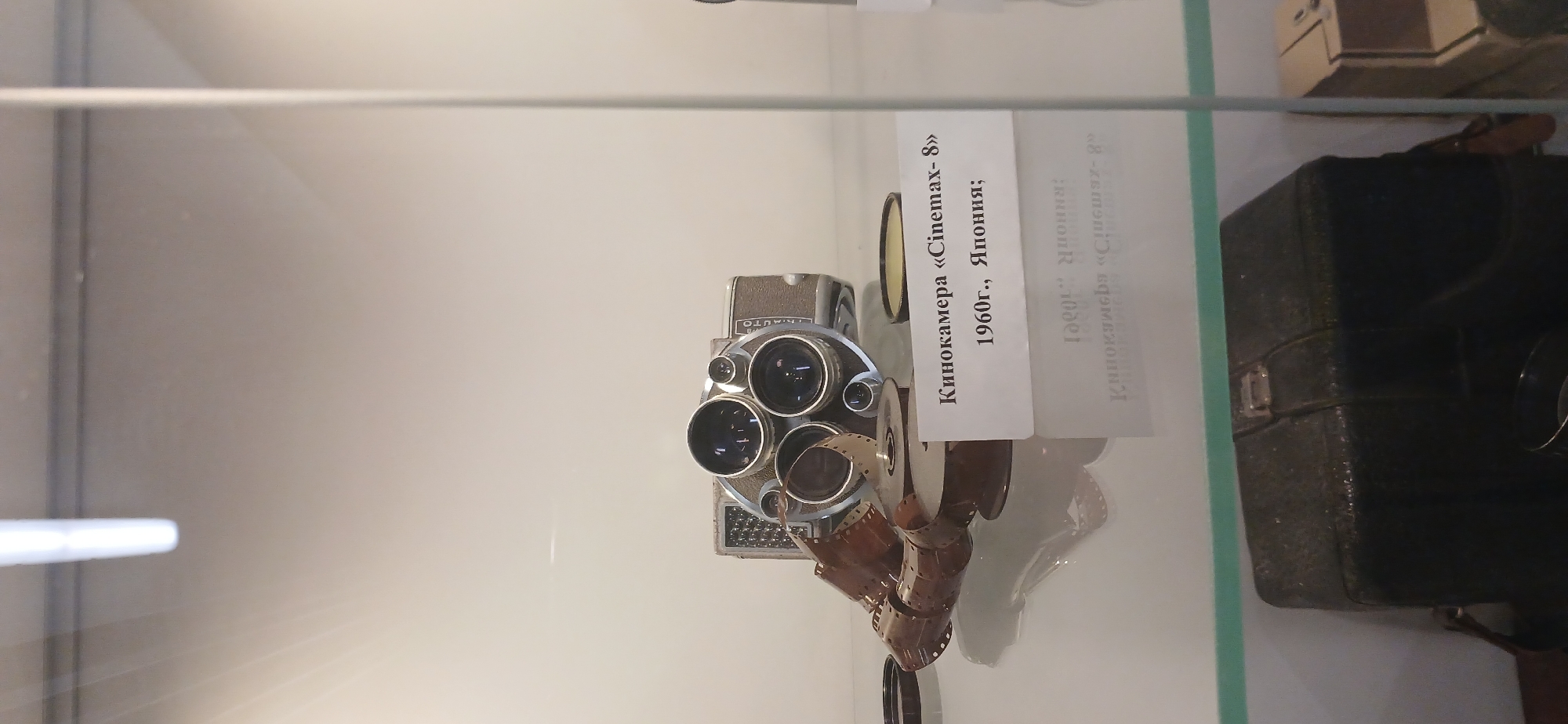 The image displays a vintage cine camera and accessories, arranged within a glass display case, likely in a museum or exhibition setting in Moscow, Russia. The photograph itself is rotated 90 degrees clockwise, so the text on the accompanying label appears vertically oriented.
The central subject is a polished metal cine camera, possibly from the 1960s, featuring a distinctive turret with three lenses of varying sizes, typical for 8mm film cameras of that era. One reel of brown film, partially unspooled and draped in front of the camera, is also visible. The camera has "AUTO" text embossed on its top.
To the right of the camera, a white descriptive label provides details in Russian. The main text reads: "Кинокамера «Cinemax- 8»" (Cine camera "Cinemax-8"), followed by "1960г., Япония;" (1960, Japan;). A clear reflection of this text, inverted and slightly distorted, is visible behind the physical label on the display surface.
Further to the right, a dark, textured case, possibly leather or a similar material, with a metal clasp, can be seen. This case likely belongs to the camera. The entire scene is enclosed within what appears to be a clear glass display, with reflections of the surroundings and the camera itself visible on the glass surfaces. The background is a neutral, light color, typical of museum exhibits.