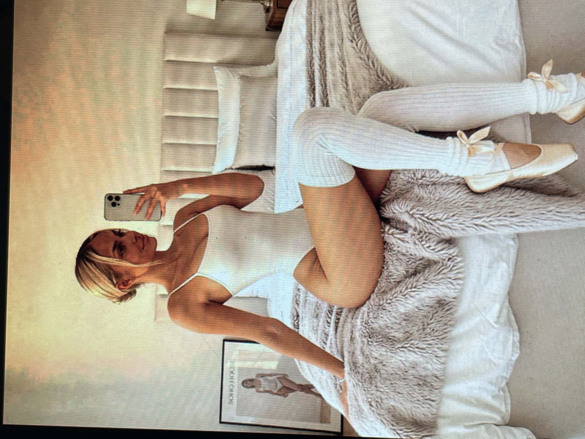 The image shows a young woman with blonde hair pulled back in a bun, wearing a white leotard and white thigh-high leg warmers tied with satin bows. She is also wearing ballet pointe shoes. She is sitting on a bed in a bedroom, taking a mirror selfie with a mobile phone. The bed has a grey, furry throw blanket and white linens. A framed picture of a woman in a leotard hangs on the wall to the left of the bed, and a headboard with vertical panels is visible behind her. A nightstand with a lamp is partially visible to the right. The room appears to be well-lit, with soft, warm lighting. The image does not contain any information that would allow for its location to be identified in the city of Mantov, Czechia.
