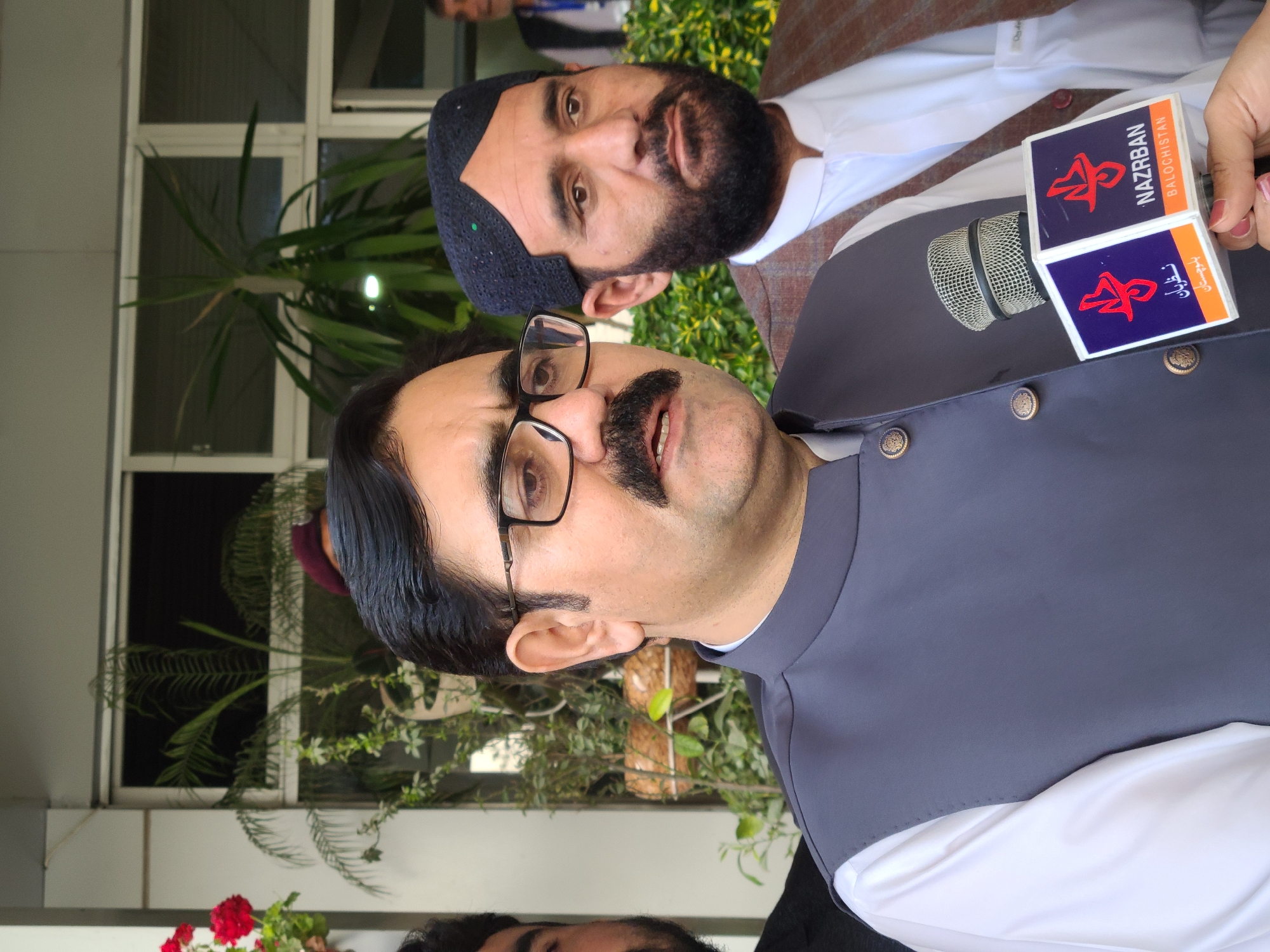 The image depicts two men in Quetta City Tehsil, Pakistan, likely during an interview. One man, in the foreground, wears glasses, a white shirt, and a grey waistcoat, addressing a microphone bearing the logo "NAZRZBAN BALOCHISTAN." The second man, behind, sports a beard and a traditional cap. The background includes a window with plants.