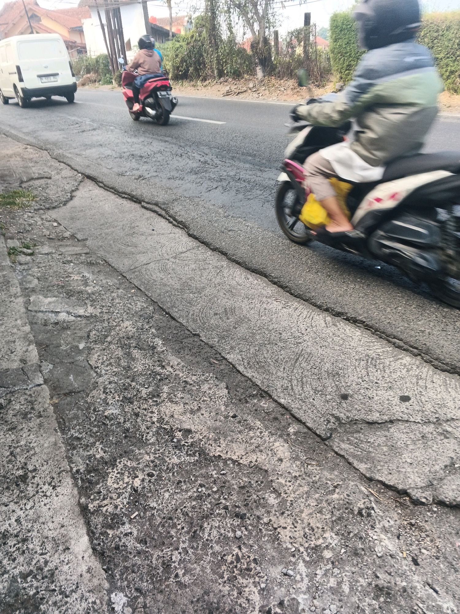 This is a motion-blurred, medium-shot, eye-level photograph taken from a sidewalk on a paved road in Ungaran, Indonesia. The road has a rough texture with visible cracks and patches. On the right side of the road, a person wearing a helmet and a gray and green jacket is riding a motorcycle, also in motion. The person's legs are visible, with one foot wearing a black sandal and the other in a yellow bag. On the left side of the road, further in the background, another person on a red motorcycle is also moving. A white van is parked on the left side of the road. The environment appears to be a semi-urban or rural area, with vegetation and buildings visible in the background. The lighting suggests it is daytime, with sunlight casting shadows.